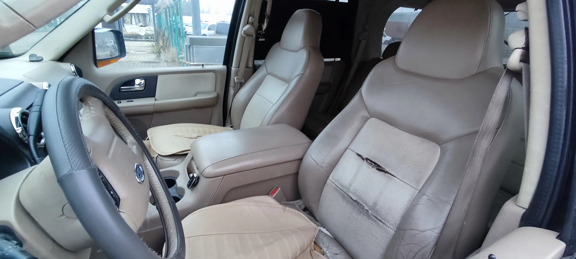 Wnętrze samochodu z beżowymi skórzany siedzeniami. Widoczne uszkodzenie w postaci rozdarcia na siedzeniu pasażera. Kierownica po lewej stronie, deska rozdzielcza i drzwi z beżowymi panelami. Otwarte drzwi pokazują zewnętrzny widok z ogrodzeniem i budynkami. W tle widoczne są inne pojazdy i zielona siatka.