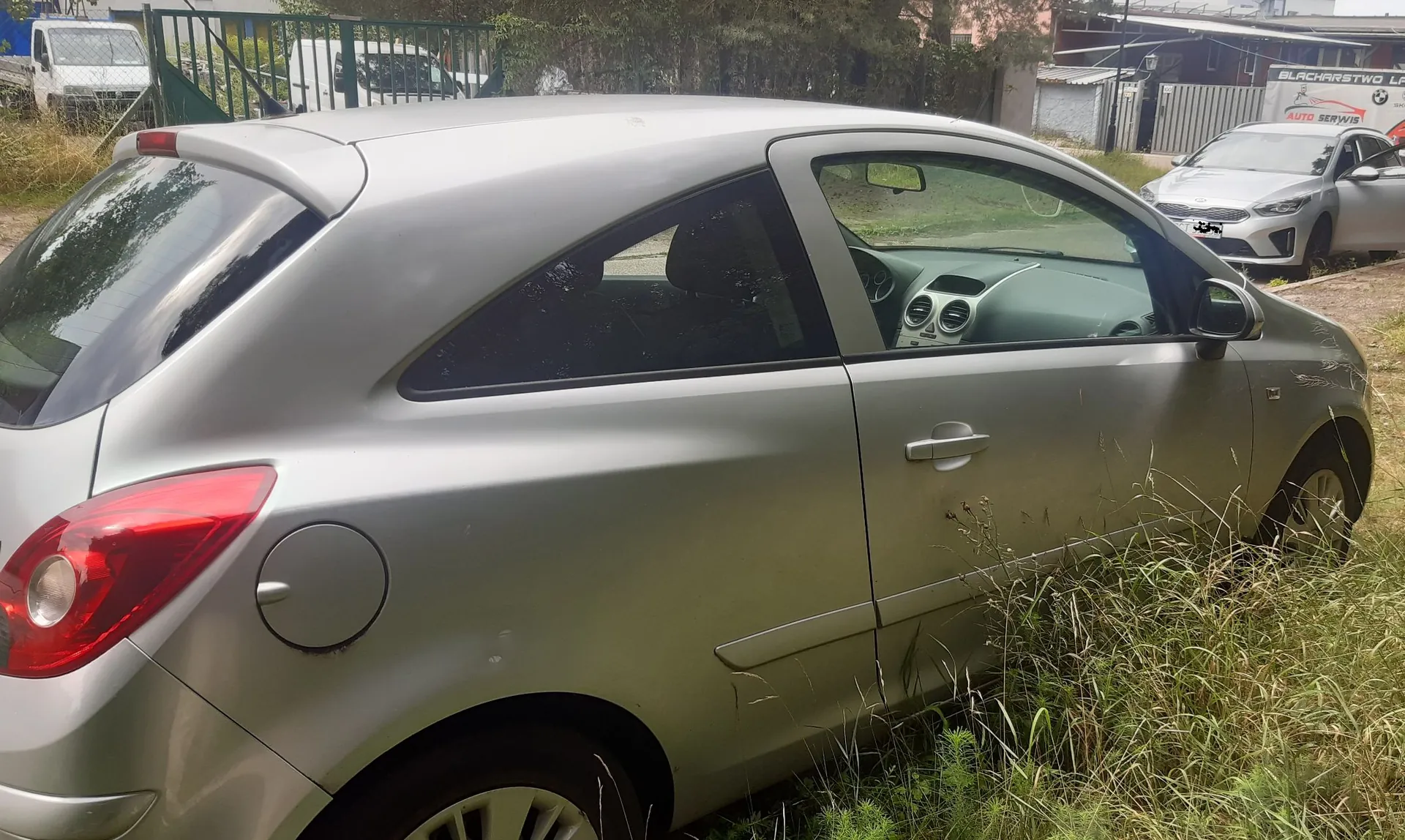 Srebrny samochód osobowy typu hatchback zaparkowany na trawiastym terenie. Widoczny jest bok auta, tylna lampa oraz wlew paliwa. W tle widać biały samochód, budynek z napisem 'BLACHARSTWO' oraz 'AUTO SERWIS' i zielone drzewa.