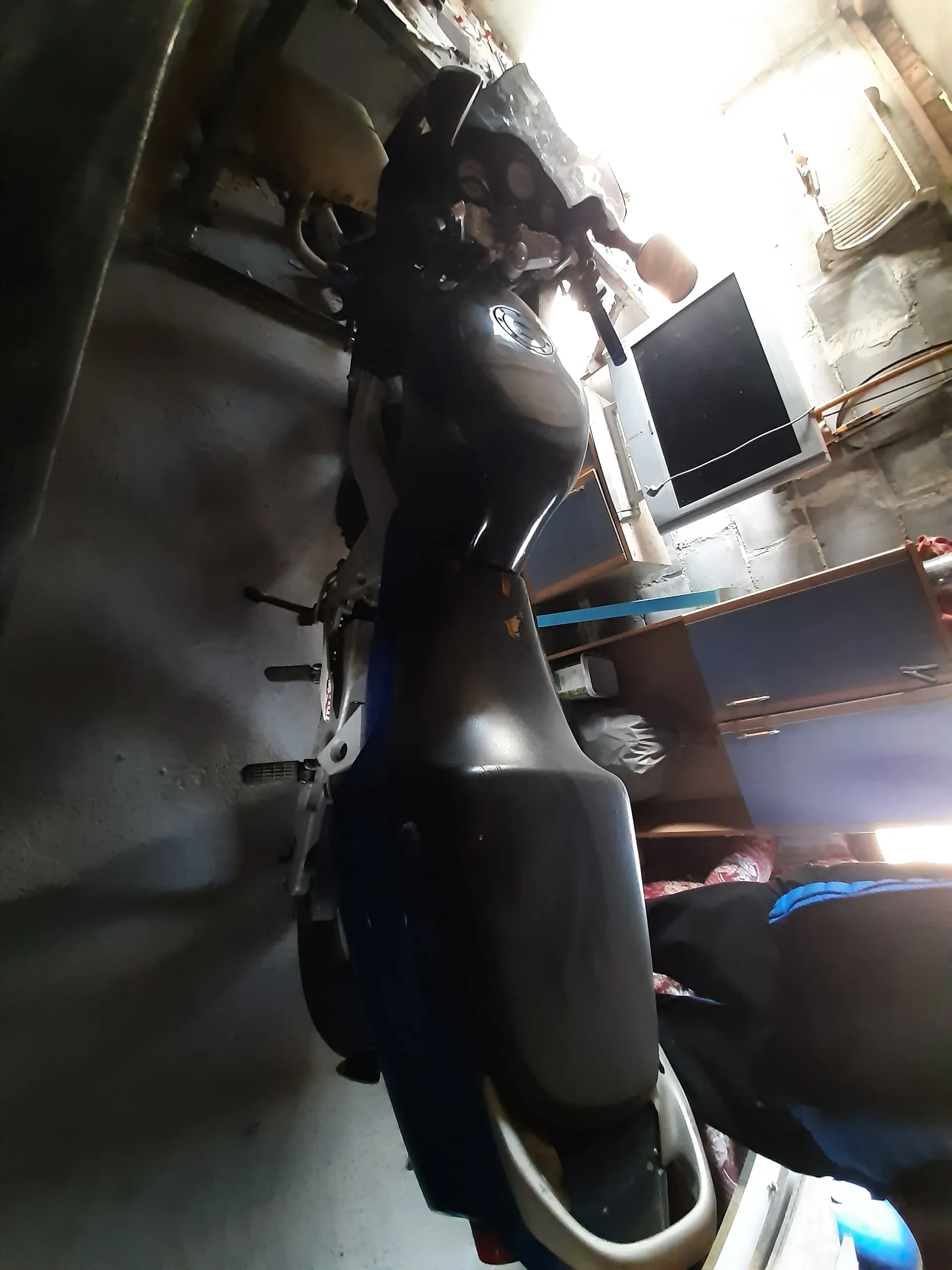 Motocykl czarny z niebieskimi elementami stojący na podstawce w garażu. W tle betonowa ściana, stary telewizor kineskopowy na biurku, niebieska szafka oraz osoba w ciemnych spodniach i niebieskiej koszuli. Oświetlenie pochodzi z okna po lewej stronie.