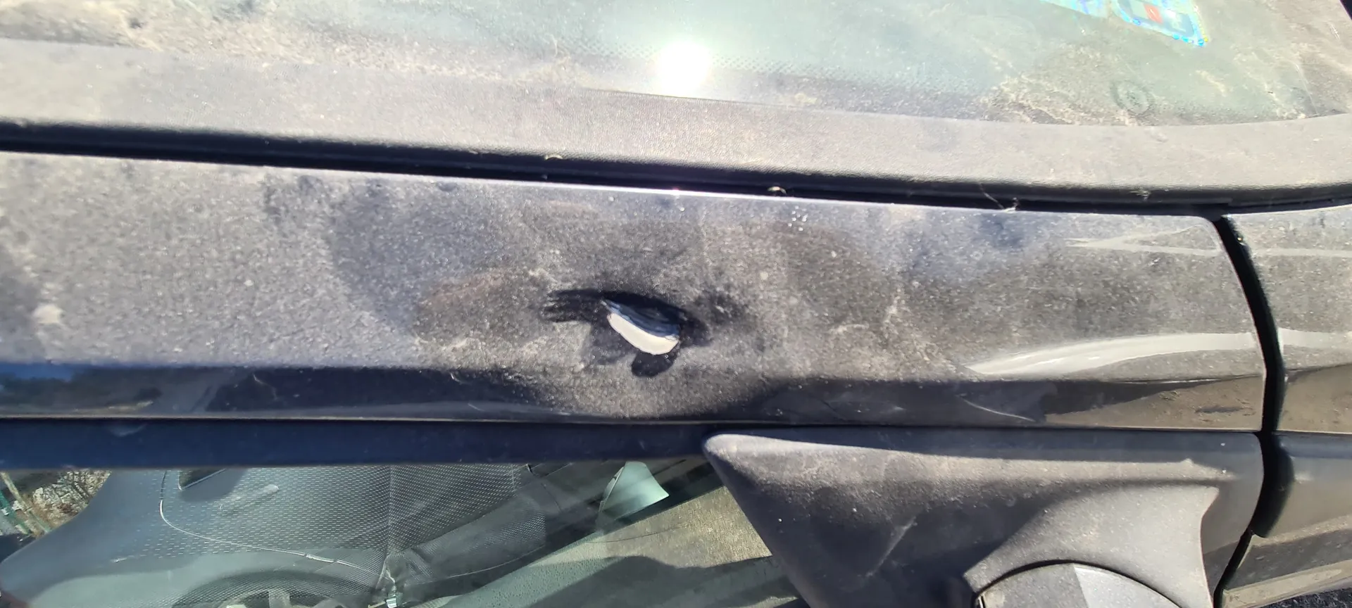 Zbliżenie na czarną plastikową obudowę przedniej części samochodu, prawdopodobnie nad kierunkowskazem. Na powierzchni widoczne jest uszkodzenie w postaci otworu z białym materiałem wewnątrz. Obudowa pokryta jest kurzem i zabrudzeniami. W tle fragment przedniej szyby z częściowo widocznym kolorowym naklejką oraz odbiciem otoczenia. Na szybie widoczne są ślady brudu i plamy.