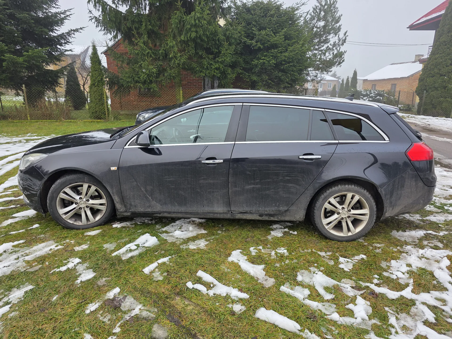 Ciemnoszary samochód typu kombi zaparkowany na trawie z resztkami śniegu. W tle widoczne są drzewa iglaste, ceglany dom oraz inne budynki z śniegiem na dachach. Na trawie i drodze po prawej stronie znajdują się plamy śniegu. Niebo jest zachmurzone. Za głównym pojazdem stoi inny samochód.