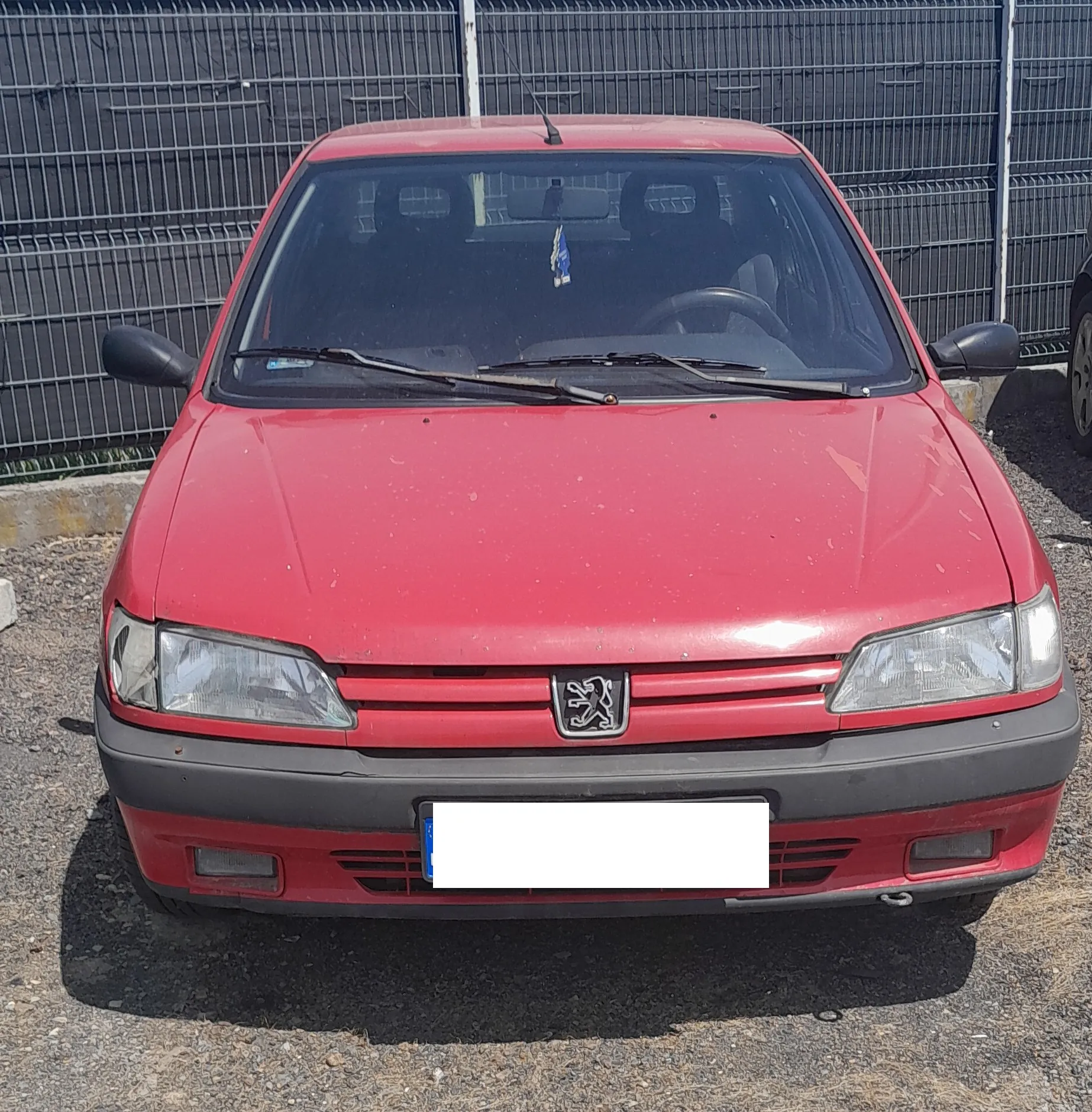 Widok z przodu czerwonego samochodu marki Peugeot, zaparkowanego na żwirowym podłożu przed szarym ogrodzeniem z metalowej siatki. Samochód ma czarny zderzak z białą, zakrytą tablicą rejestracyjną oraz charakterystyczne logo lwa na grillu.