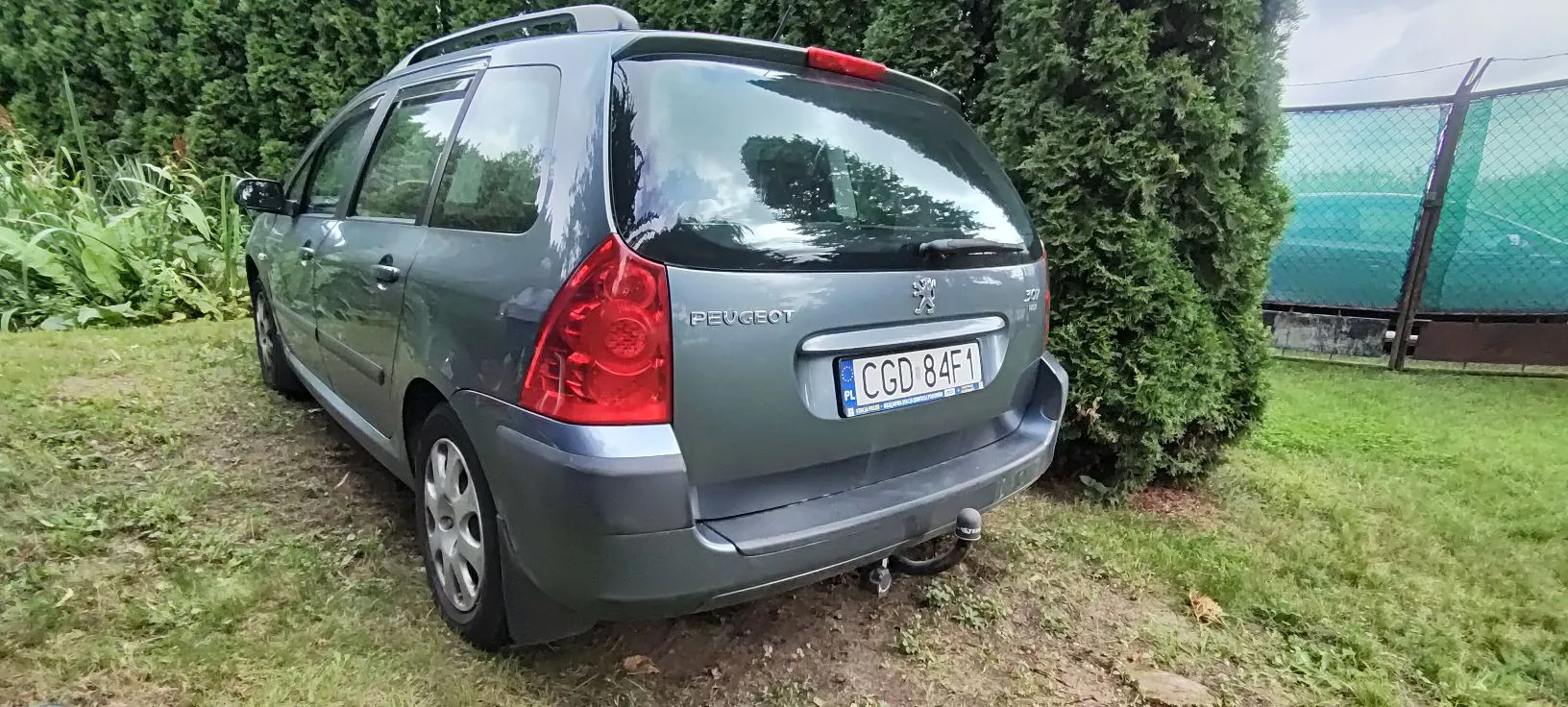 Szary samochód marki Peugeot w nadwoziu kombi, zaparkowany na trawie przed zielonymi żywopłotami. Widoczny jest tył pojazdu z tablicą rejestracyjną o numerze CGD 84F1, logo marki oraz napisem PEUGEOT. Samochód posiada relingi dachowe i hak holowniczy.