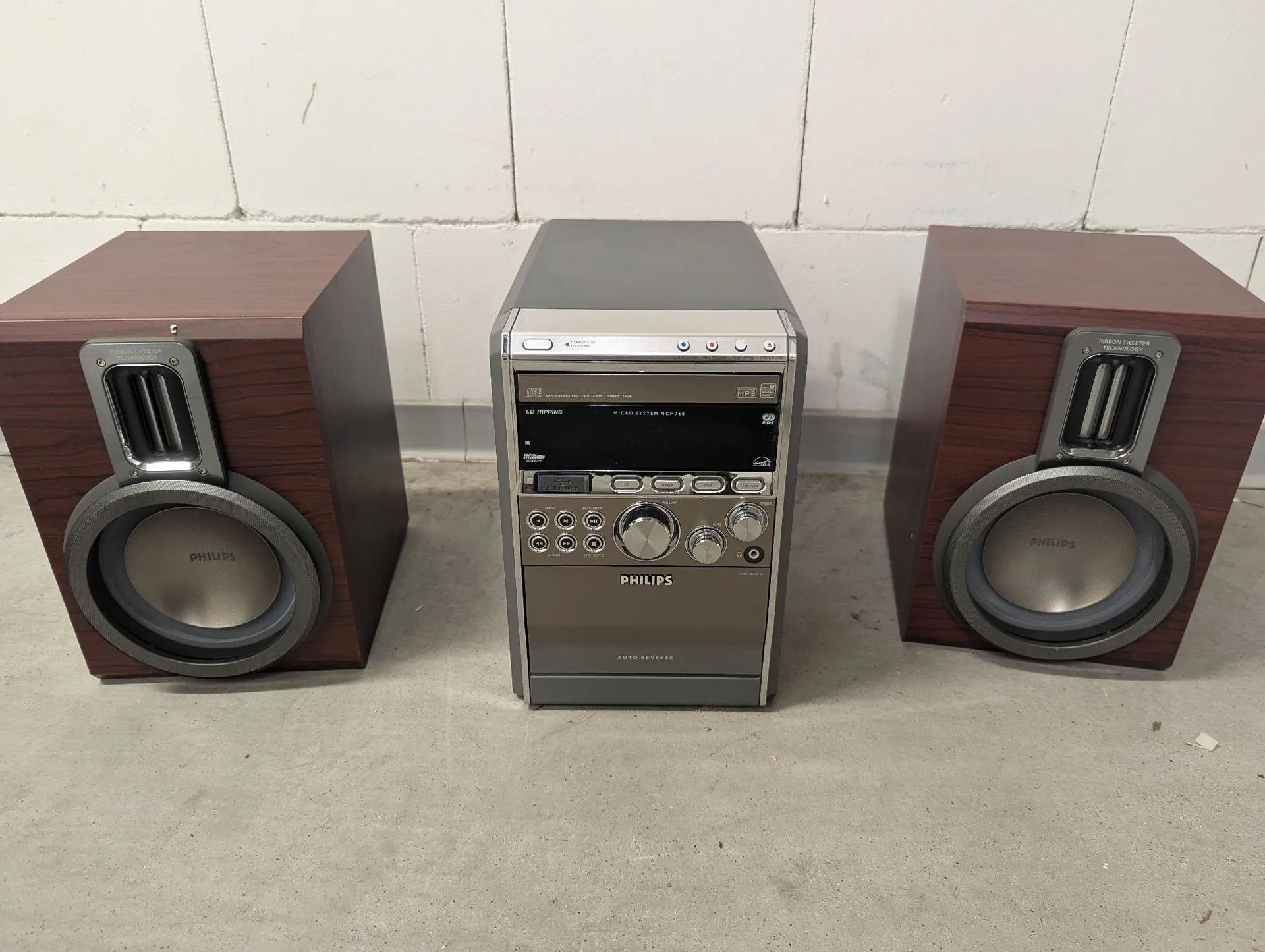 Zestaw audio marki Philips składający się z centralnej jednostki sterującej oraz dwóch głośników satelitów w obudowach o kolorze ciemnego drewna. Jednostka centralna jest srebrna, posiada wyświetlacz, pokrętła, przyciski sterujące oraz napis 'PHILIPS' i 'AUTO REVERSE'. Głośniki mają srebrne membrany z logo 'PHILIPS' oraz górne otwory basowe. Całość stoi na jasnej, betonowej podłodze na tle białej ściany.