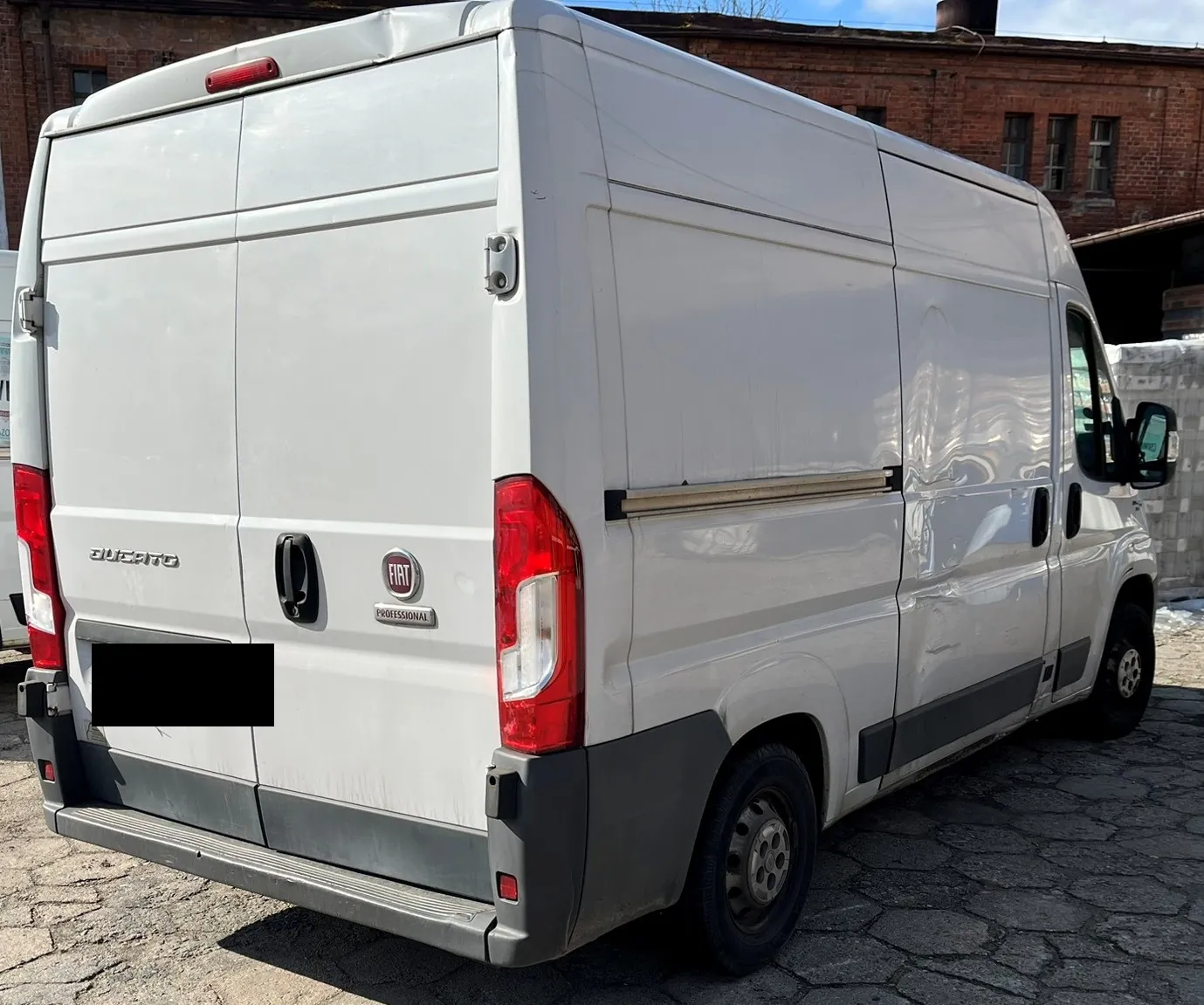 Biały furgon marki Fiat Ducato zaparkowany na kostce brukowej na tle ceglanego budynku. Pojazd jest widoczny od tyłu i z boku. Na tylnych drzwiach widnieją napisy 'DUCATO' oraz logo 'FIAT PROFESSIONAL'. Prawa strona nadwozia samochodu jest wyraźnie wgnieciona i uszkodzona. Tablica rejestracyjna jest zakryta czarnym polem.