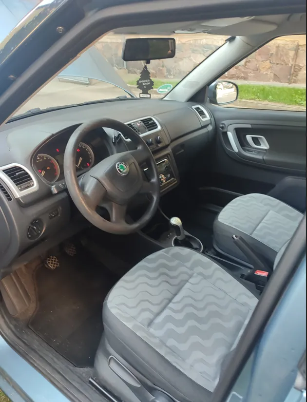 Wnętrze samochodu marki Škoda widziane z perspektywy otwartych drzwi pasażera. Widoczna jest ciemnoszara deska rozdzielcza, kierownica z logo Škoda, zegary, konsola środkowa z radiem oraz dwa przednie fotele z szarym obiciem w poziome, faliste wzory. W lusterku wstecznym wisi zapach samochodowy w kształcie choinki z napisem 'WUNDER BAUM'.