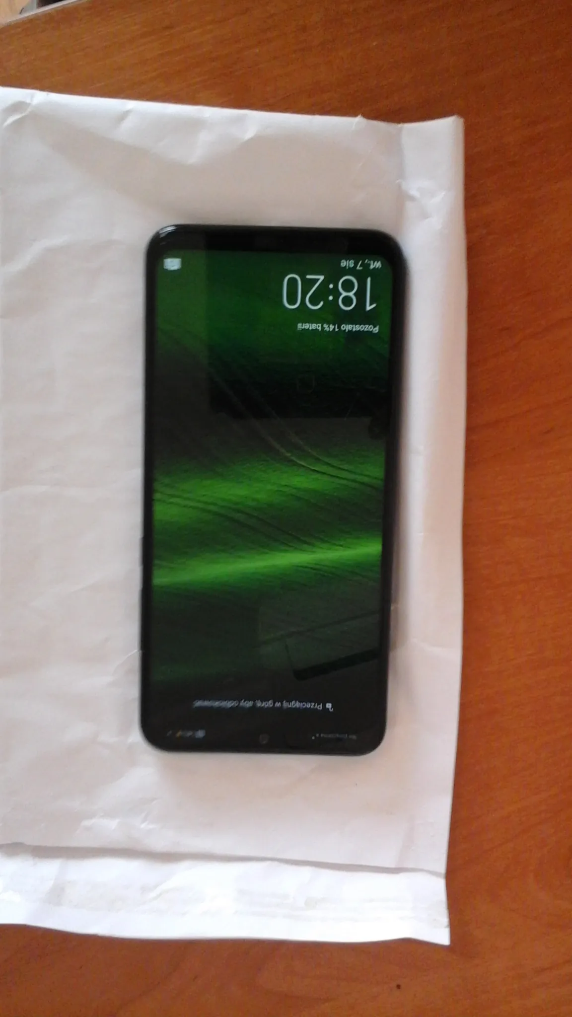 Smartfon leżący na białym papierze na drewnianym stole. Ekran wyświetla zieloną tapetę przypominającą zorzę polarną. Na ekranie widoczny czas 18:20, napis 'Pozostało 14% baterii', data 'Wt, 7 sie' oraz instrukcja 'Przesuń w górę, aby odblokować'.