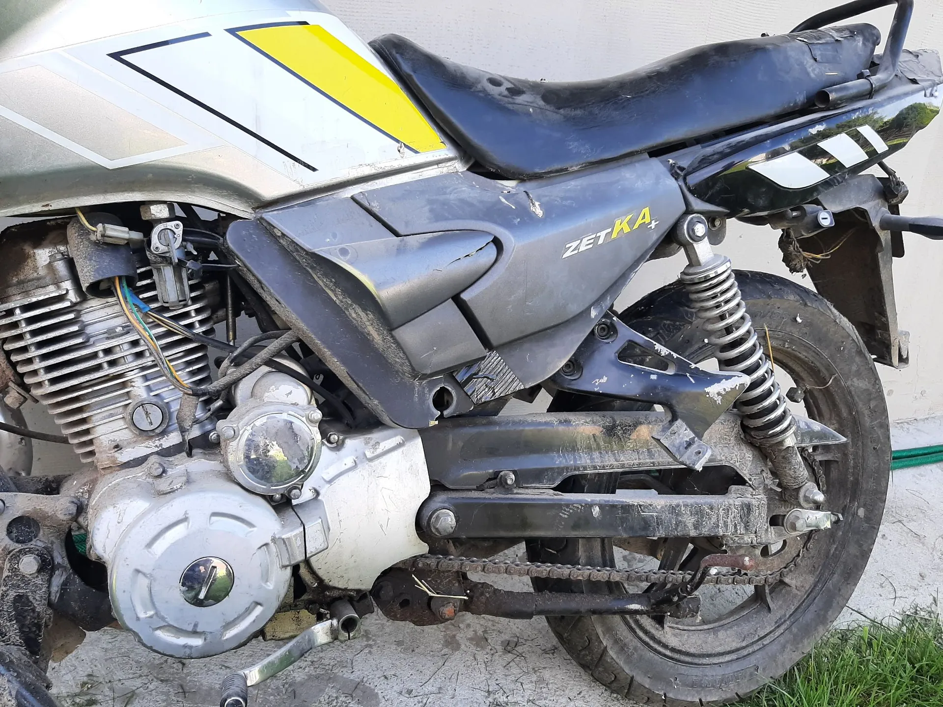 Zbliżenie na prawą stronę motocykla w kolorze srebrnym z żółtym i czarnym pasem na baku. Widoczny jest czarny siedzenie, silnik, łańcuch napędowy oraz tylne koło z amortyzatorem. Na czarnym elemencie nad tylnym kołem znajduje się biały napis „ZETKA”.
