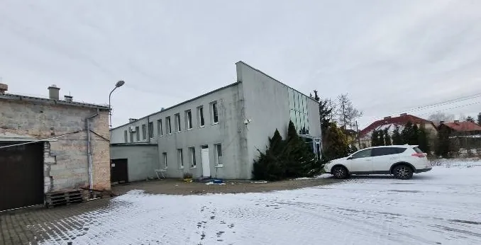 Zdjęcie przedstawia zimowy krajobraz z budynkami. W centrum znajduje się szary, dwupiętrowy budynek z betonowymi ścianami i prostokątnymi oknami. Po lewej stronie widoczny jest starszy, ceglany budynek z kominem i zamkniętą bramą. Przy budynku stoi latarnia uliczna. Na prawo od głównego budynku zaparkowany jest biały SUV. Teren pokryty jest śniegiem z widocznymi śladami stóp. W tle znajdują się domy z czerwonymi dachami oraz drzewa. Niebo jest zachmurzone.