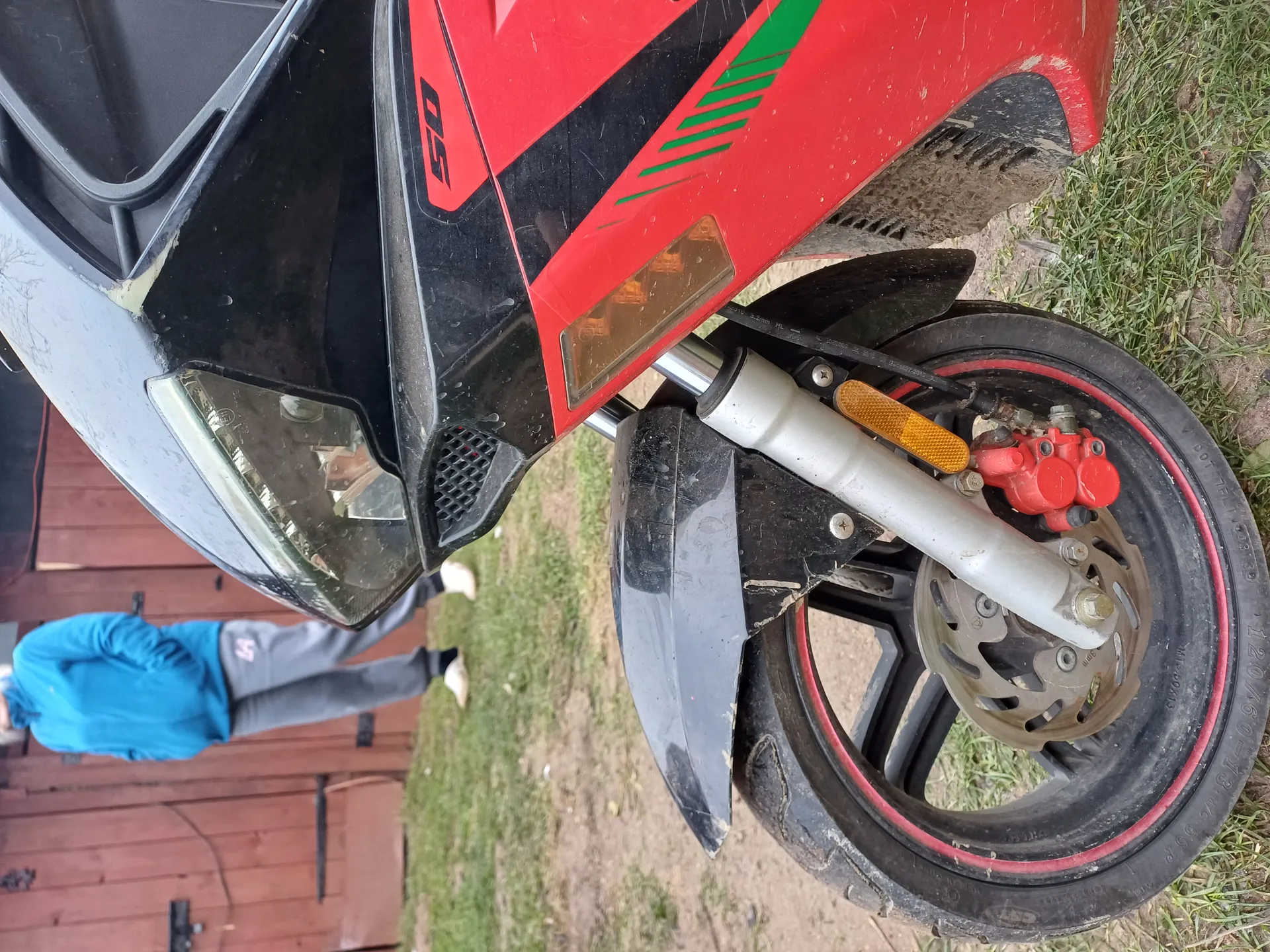 Czerwono-czarny skuter z napisem "50", czarną osłoną przednią, reflektorem, żółtym reflektorem kierunkowskazu i przednim kołem z czerwonymi akcentami na obręczy oraz czerwonymi hamulcami. Osoba w niebieskiej kurtce i szarych spodniach stoi na trawie obok drewnianego ogrodzenia. Skuter lekko zabrudzony kurzem.