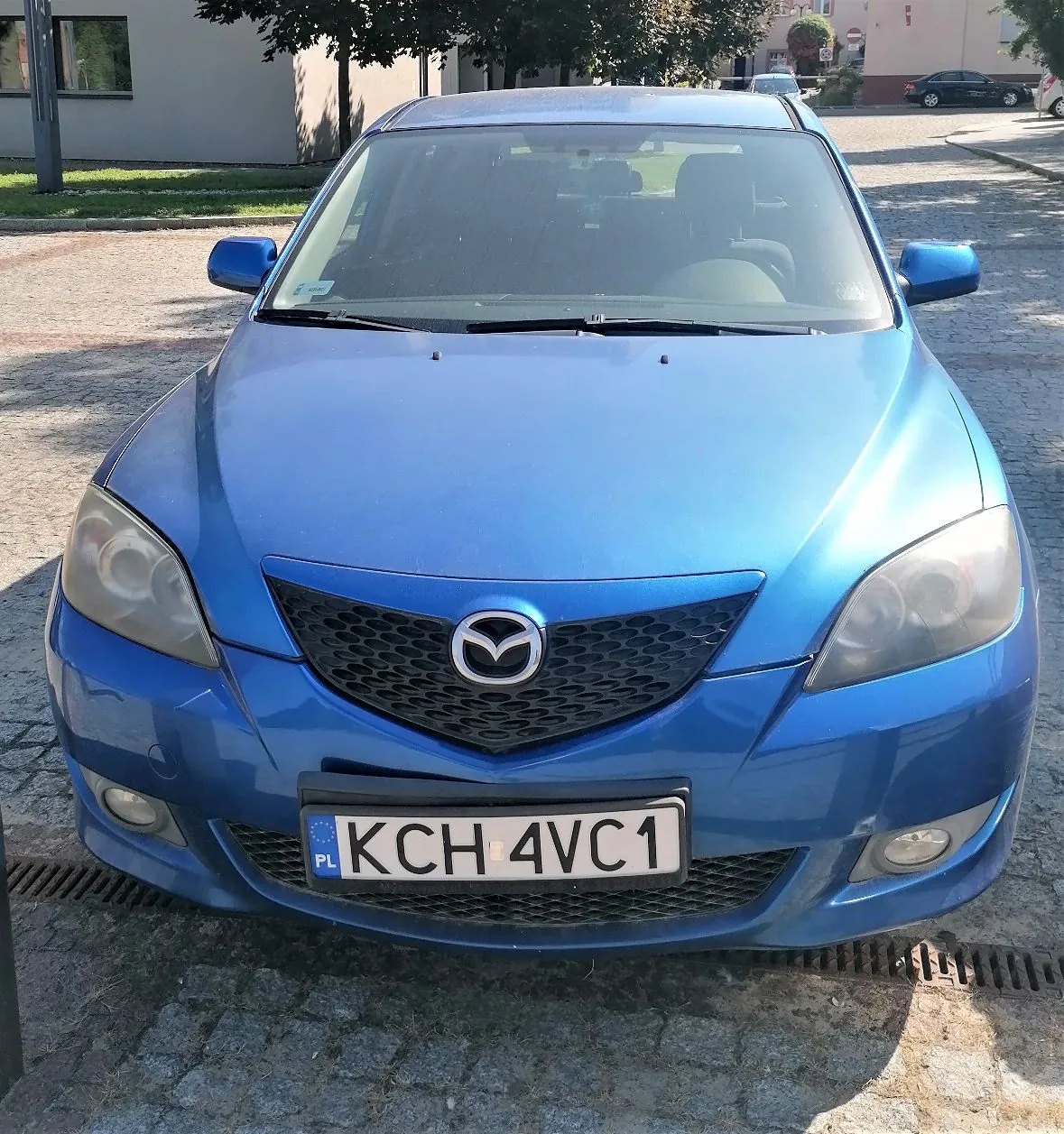 Niebieski samochód marki Mazda zaparkowany przodem do obiektywu na kostce brukowej. Widoczna jest przednia część pojazdu z grillem, logo Mazdy oraz polską tablicą rejestracyjną o numerze KCH 4VC1.