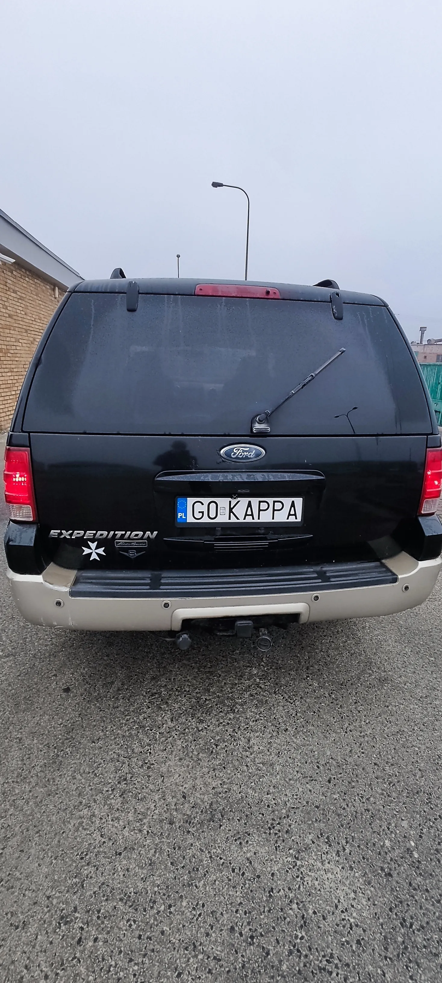 Tyl czarnego samochodu Ford Expedition zaparkowanego na asfalcie. Widoczna tablica rejestracyjna z napisem GO KAPPA oraz niebieskim pasem z oznaczeniem PL. Na tyle samochodu znajduje się logo Ford, napis EXPEDITION oraz biała naklejka w kształcie gwiazdy. W tle widać fragment budynku z cegły, latarnię uliczną oraz pochmurne niebo. Samochód jest ustawiony pionowo, co sugeruje nieprawidłową orientację obrazu.