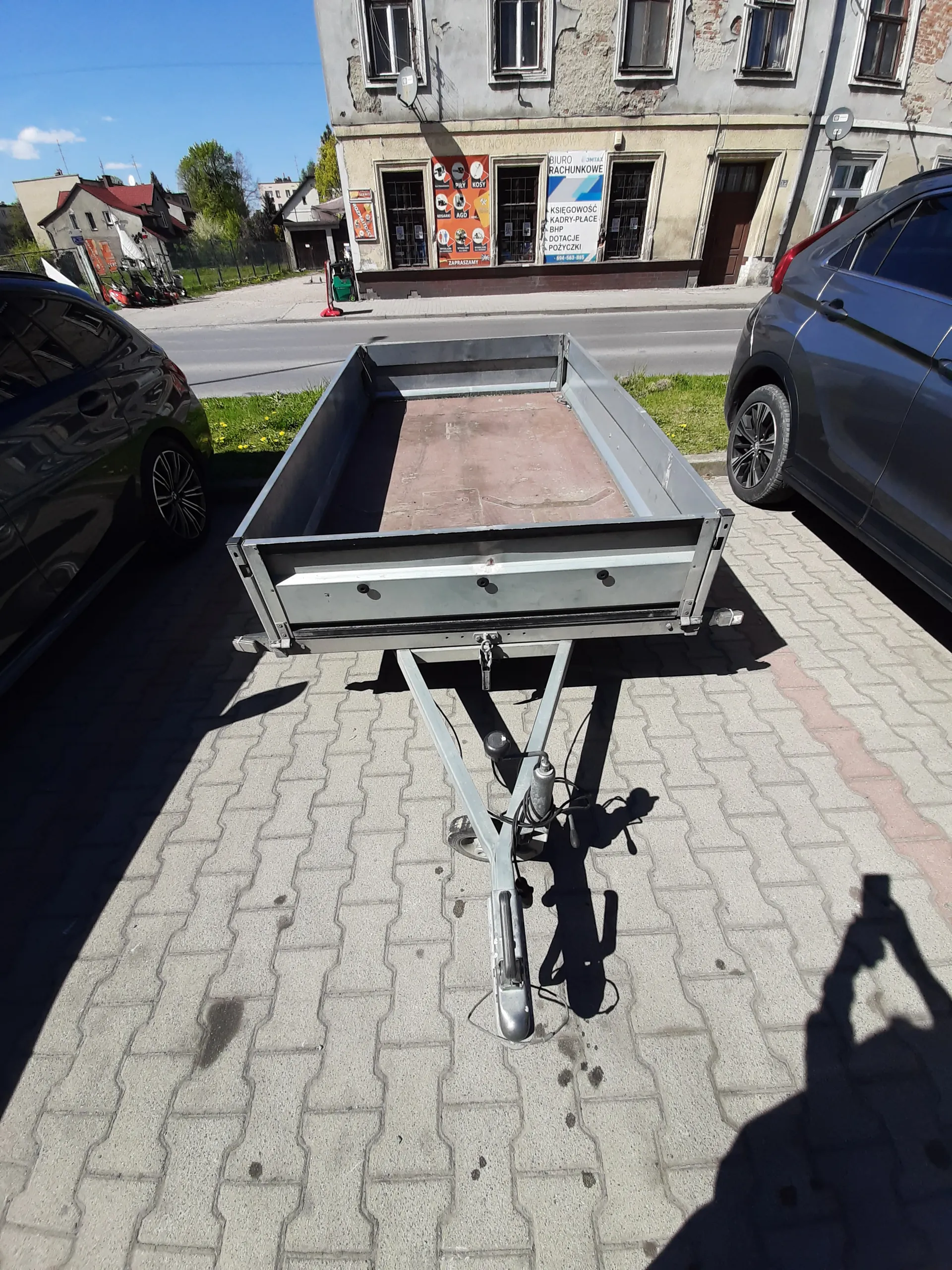 Metalowa przyczepa na brukowanej powierzchni, dwa samochody (ciemnoniebieski i czarny), budynek z oknami i szyldami. Szyldy zawierają tekst: RACHUNKOWE, KSIĘGOWOŚĆ, POŻYCZKI, BHP, DOTACJE oraz pomarańczowe szyldy z napisami KAWA, HERBATA, PIW, SOKI. Widoczne cienie osób i obiektów, niebo w tle.