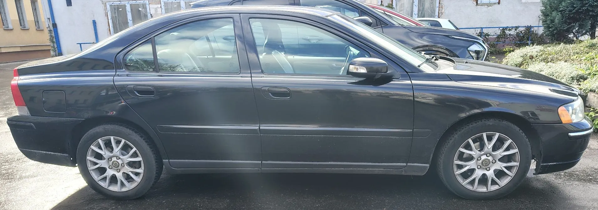 Czarny samochód osobowy typu sedan zaparkowany bokiem na ciemnym podłożu. Pojazd ma srebrne wieloramienne felgi aluminiowe. W tle widoczny jest fragment innego, ciemnego samochodu oraz jasna ściana budynku i zielone krzewy.