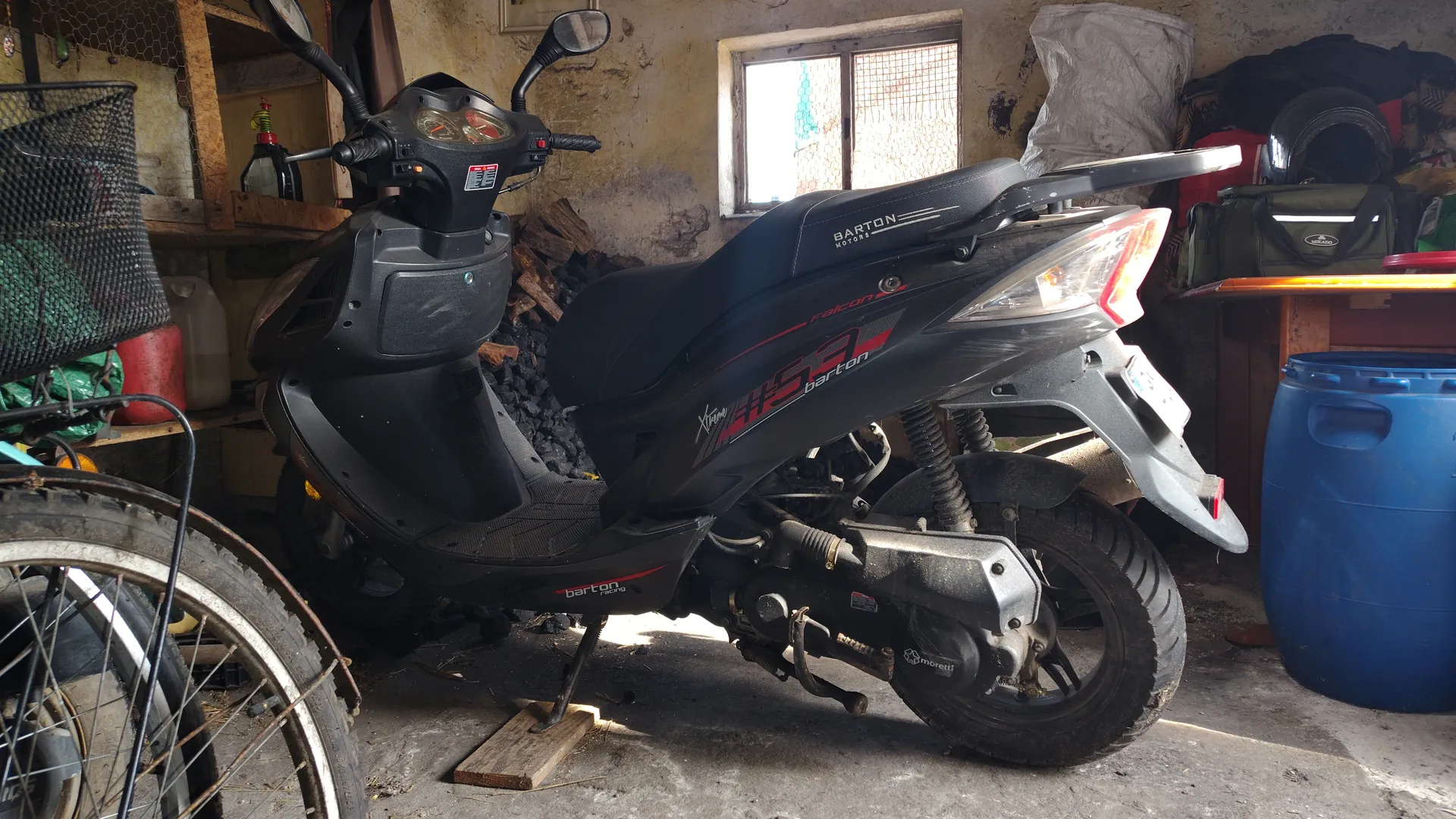Czarny skuter marki Barton z napisami 'Falcon', 'Xtreme', '150' i 'barton' na nadwoziu, zaparkowany w garażu lub warsztacie. Skuter oparty na podstawce z drewnianym klockiem pod kołem. W tle widoczne: okno z siatką, stos drewna lub węgla, niebieski beczka, torby, kask na stole oraz fragment koła roweru z siatką po lewej stronie. Ściany pomieszczenia są zużyte, z obłupioną farbą. Tekst na skuterze czytelny, ustawiony poziomo.