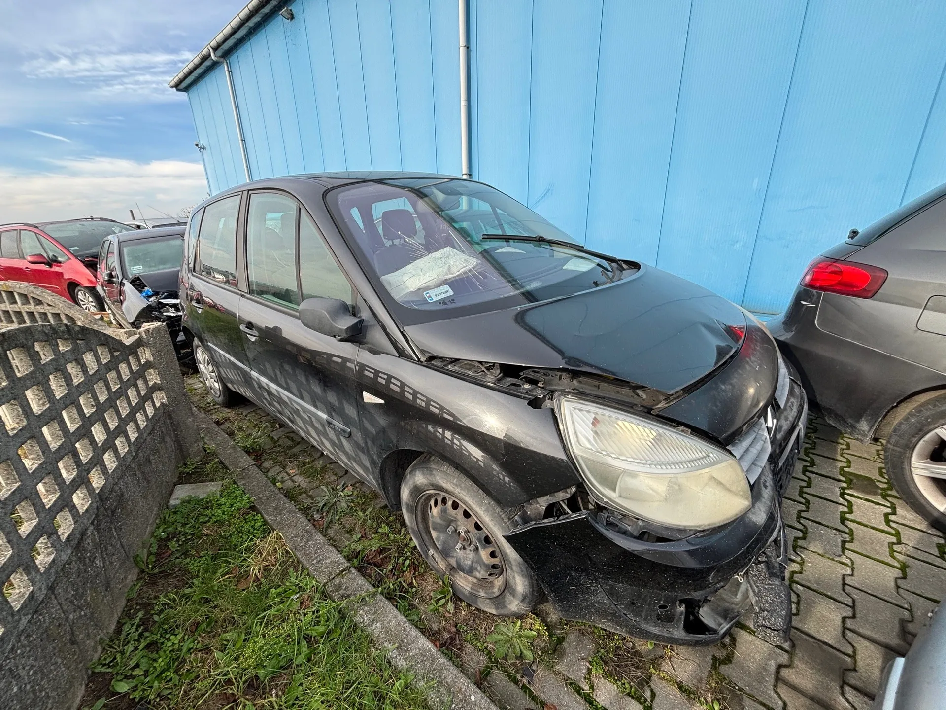 Zniszczony czarny samochód osobowy z uszkodzonym przodem, w tym zdeformowaną maską i zniszczonym reflektorem. Po lewej stronie widoczny betonowy płot z siatką, a w tle niebieski budynek przemysłowy. Wokół znajdują się inne pojazdy: czerwony i szary samochód. Podłoga składa się z chodnikowych płyt i trawy. Na niebie widoczne są chmury. Na przedniej szybie czarnego samochodu znajduje się naklejka.