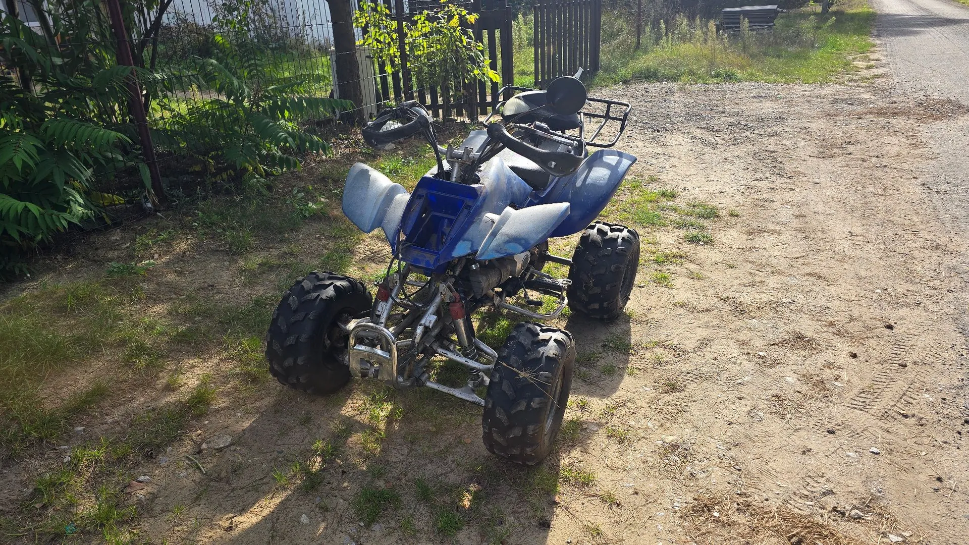 Niebieski quad terenowy zaparkowany na piaszczystej drodze obok trawnika i ogrodzenia. Pojazd jest zabrudzony błotem, posiada cztery szerokie opony z wyraźnym bieżnikiem oraz metalowy bagażnik z tyłu.