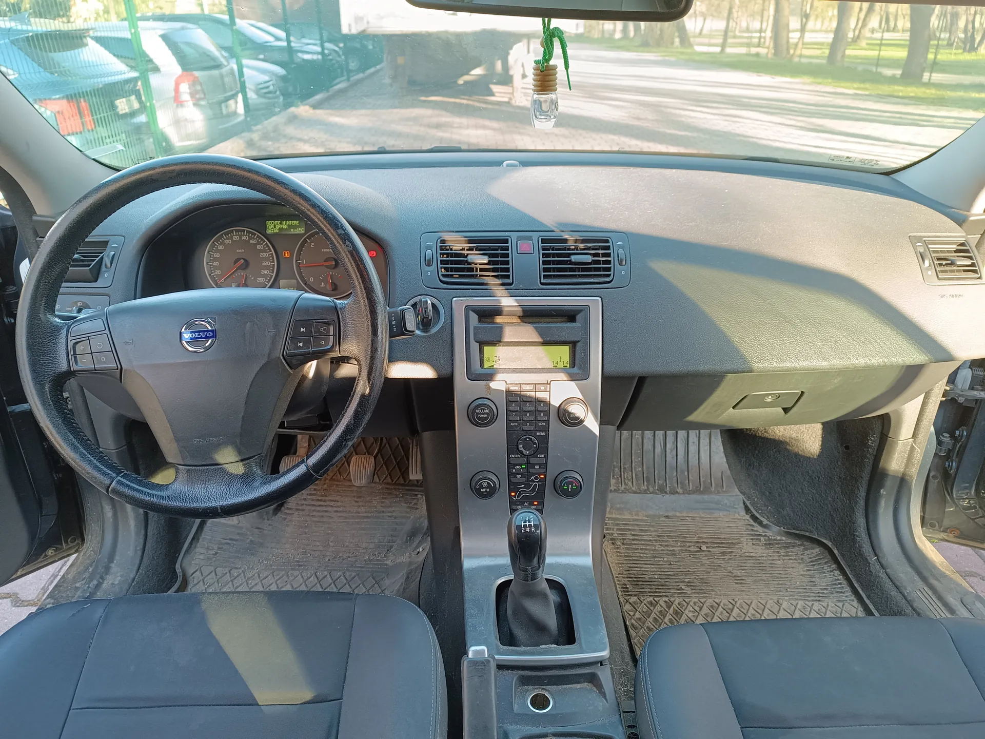 Wnętrze samochodu Volvo z widokiem na deskę rozdzielczą, kierownicę z logo marki, liczniki prędkości i obrotomierze, centralną konsolę z przyciskami i ekranem, dźwignią zmiany biegów oraz czarnymi siedzeniami. Przez przednią szybę widoczne są zaparkowane samochody, ogrodzenie i drzewa. Na lusterku wstecznym wisząca zielona ozdoba. Drzwi samochodu są otwarte.