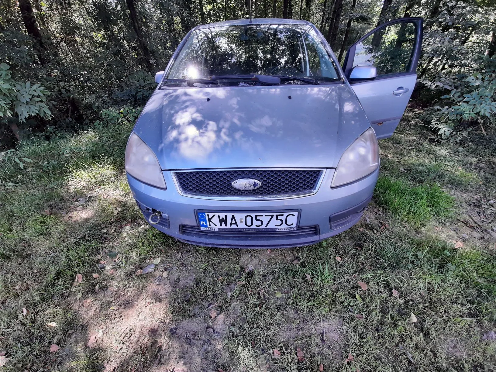Srebrny samochód marki Ford zaparkowany na trawiastym terenie w otoczeniu drzew. Pojazd ma otwarte drzwi kierowcy, a na przednim zderzaku znajduje się polska tablica rejestracyjna o numerze KWA 0575C.