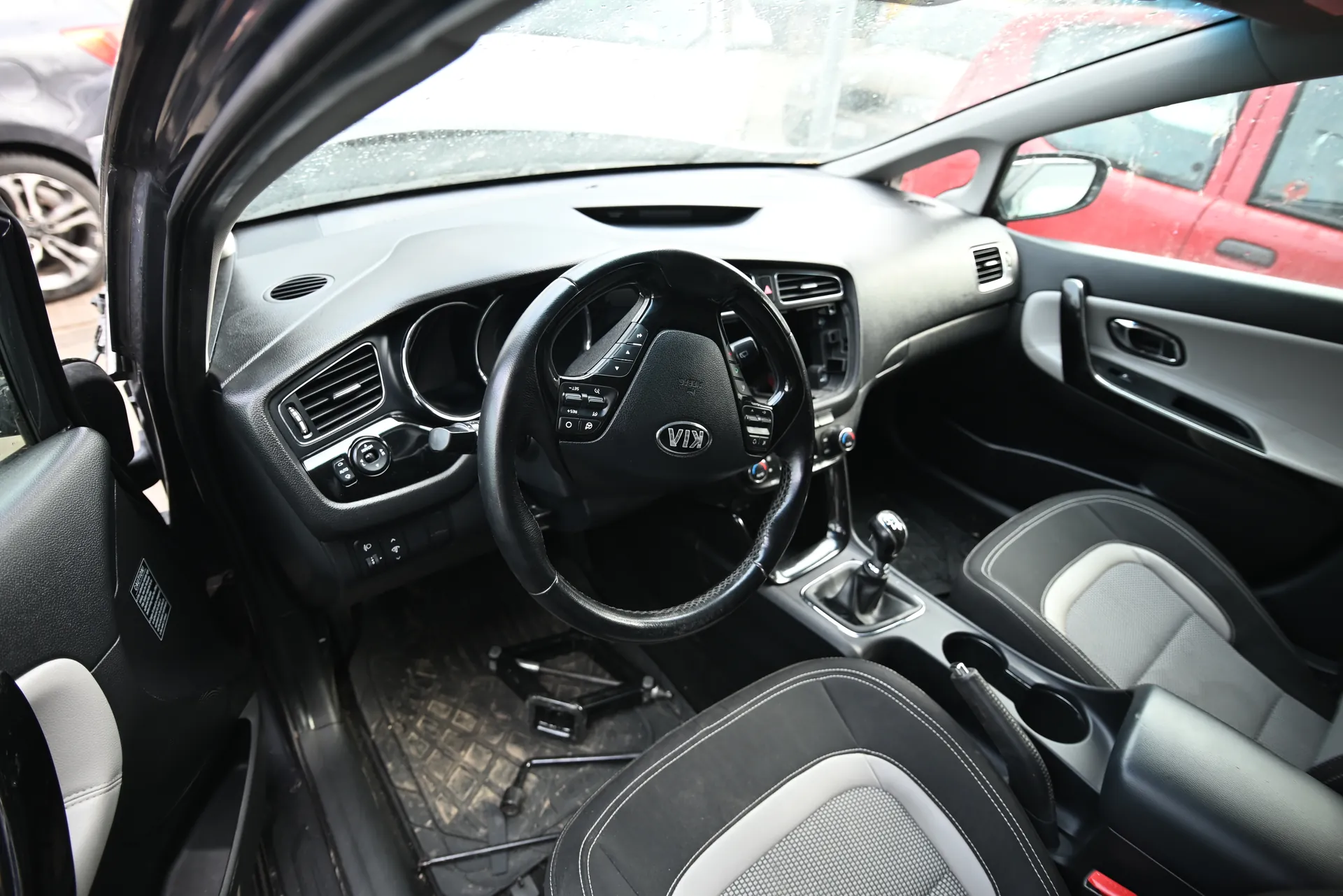 Wnętrze samochodu marki Kia z widocznym kierownicą z logo Kia, czarno-szarym deski rozdzielczej z wentylacjami i przyciskami, czarno-białymi fotelami z przeszyciami, dźwignią zmiany biegów oraz otwartą drzwiami kierowcy. Na zewnątrz przez przednią szybę widoczne są czerwony i szary samochód. Na podłodze wewnątrz pojazdu znajduje się narzędzie.