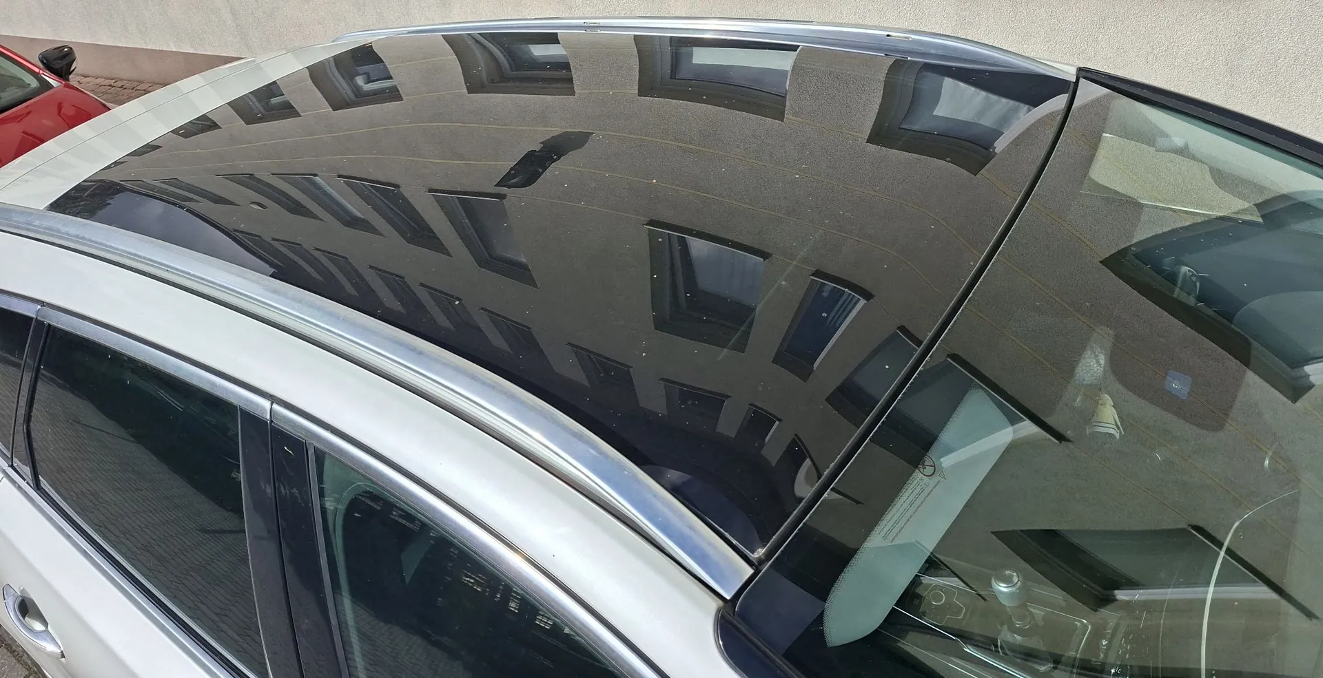 Zbliżenie na dach białego samochodu z panoramicznym, czarnym szklanym dachem. W gładkiej powierzchni szkła odbija się fasada budynku z rzędami prostokątnych okien.