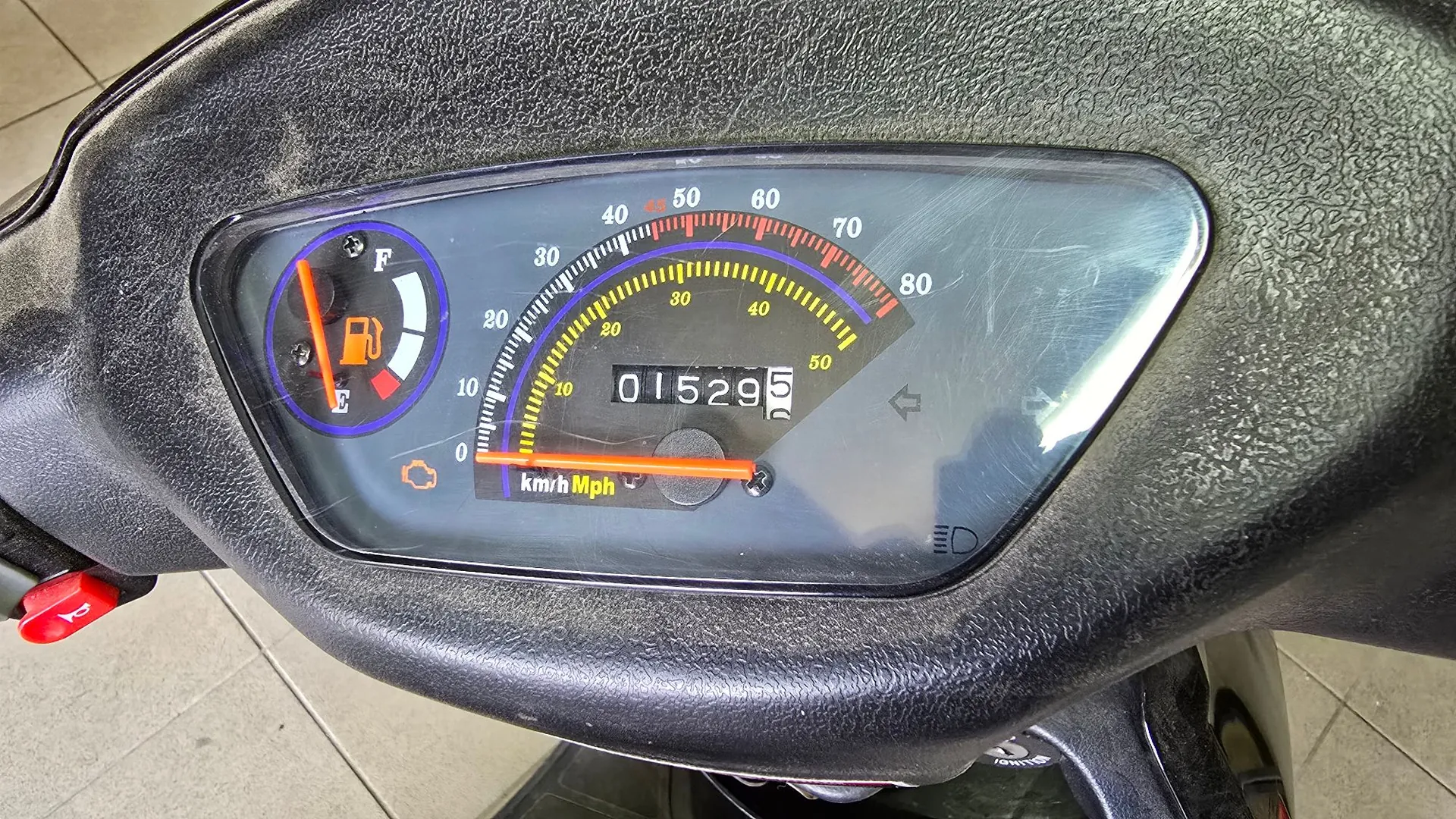 Zbliżenie na panel instrumentów skutera. Widoczny jest analogowy prędkościomierz z podziałką w km/h (do 80) i Mph (do 50). W centrum znajduje się licznik kilometrów z wartością 01529. Po lewej stronie znajduje się wskaźnik poziomu paliwa z oznaczeniami F (pełny) i E (pusty). Panel jest osadzony w czarnej, plastikowej obudowie o chropowatej teksturze.