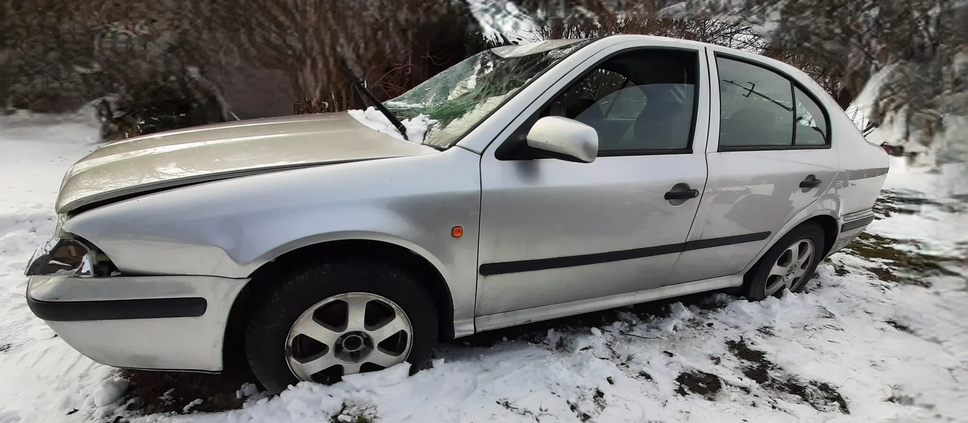 Srebrny samochód osobowy typu sedan z uszkodzonym przodem, w tym zgniecioną maską i uszkodzonym reflektorem. Na masce i przedniej szybie widoczny śnieg. Samochód stoi na śnieżnej powierzchni z widocznymi plamami ziemi. W tle drzewa i krzewy pokryte śniegiem. Pojazd ma czarne pasy dekoracyjne oraz srebrne felgi.
