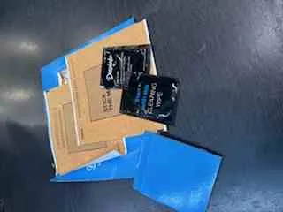 Dwa czarne opakowania chipsów Doritos Cool Ranch, brązowe pudełko kartonowe z widocznym napisem 'USPS' oraz niebieskie koperty leżące na ciemnej powierzchni.