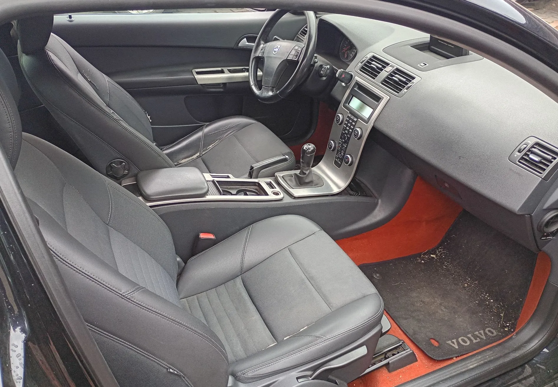 Wnętrze samochodu marki Volvo z widokiem na przednie fotele, deskę rozdzielczą i konsolę środkową. Fotele są wykonane z czarnej skóry. Widoczna jest kierownica z logo Volvo, system audio, lewarek zmiany biegów oraz pomarańczowe wykończenie podłogi pod czarną wycieraczką z napisem 'VOLVO'.