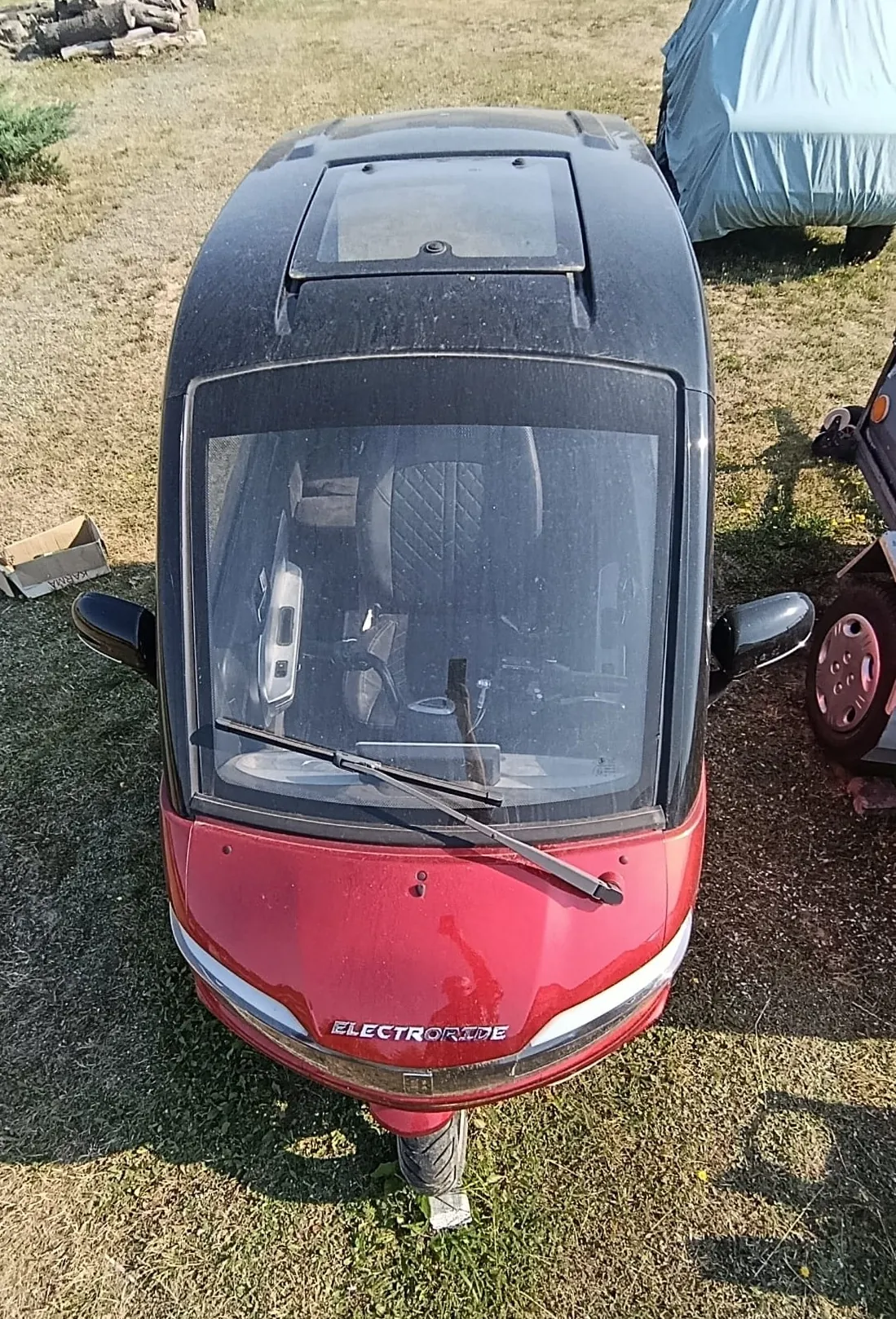 Widok z przodu na mały, czerwony pojazd elektryczny marki ElectroRide z czarnym dachem i szybą przednią. Pojazd stoi na trawiastym terenie. Na masce widnieje biały napis 'ELECTRORIDE'. Wewnątrz widoczne są ciemne fotele z przeszyciami w romby.