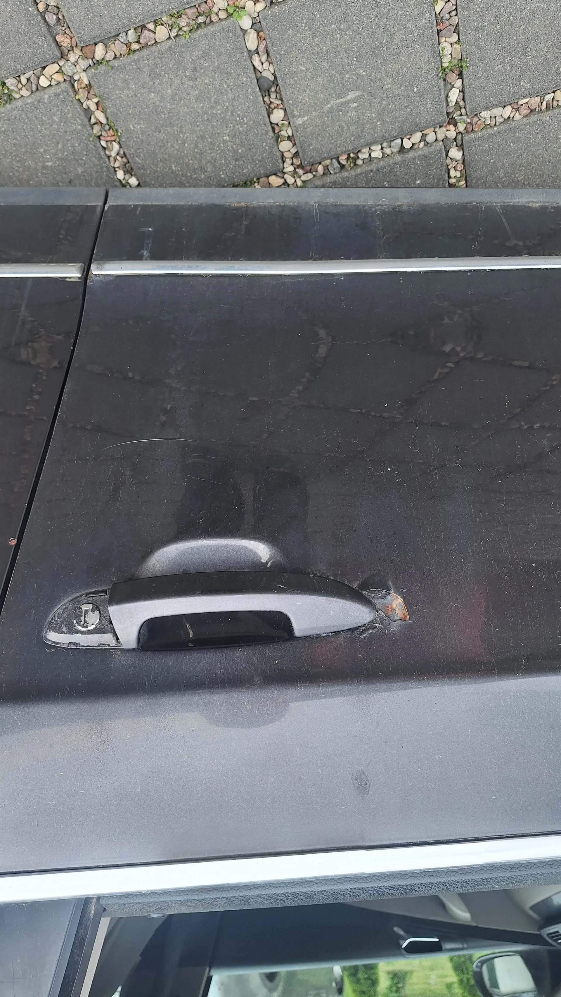 Zbliżenie na czarną drzwi samochodu z pionową klamką. W okolicy klamki widoczne uszkodzenie z oznakami rdzy. W tle chodnik z kamiennymi płytami i widok na wnętrze otwartego drzwi z lusterkiem bocznym.