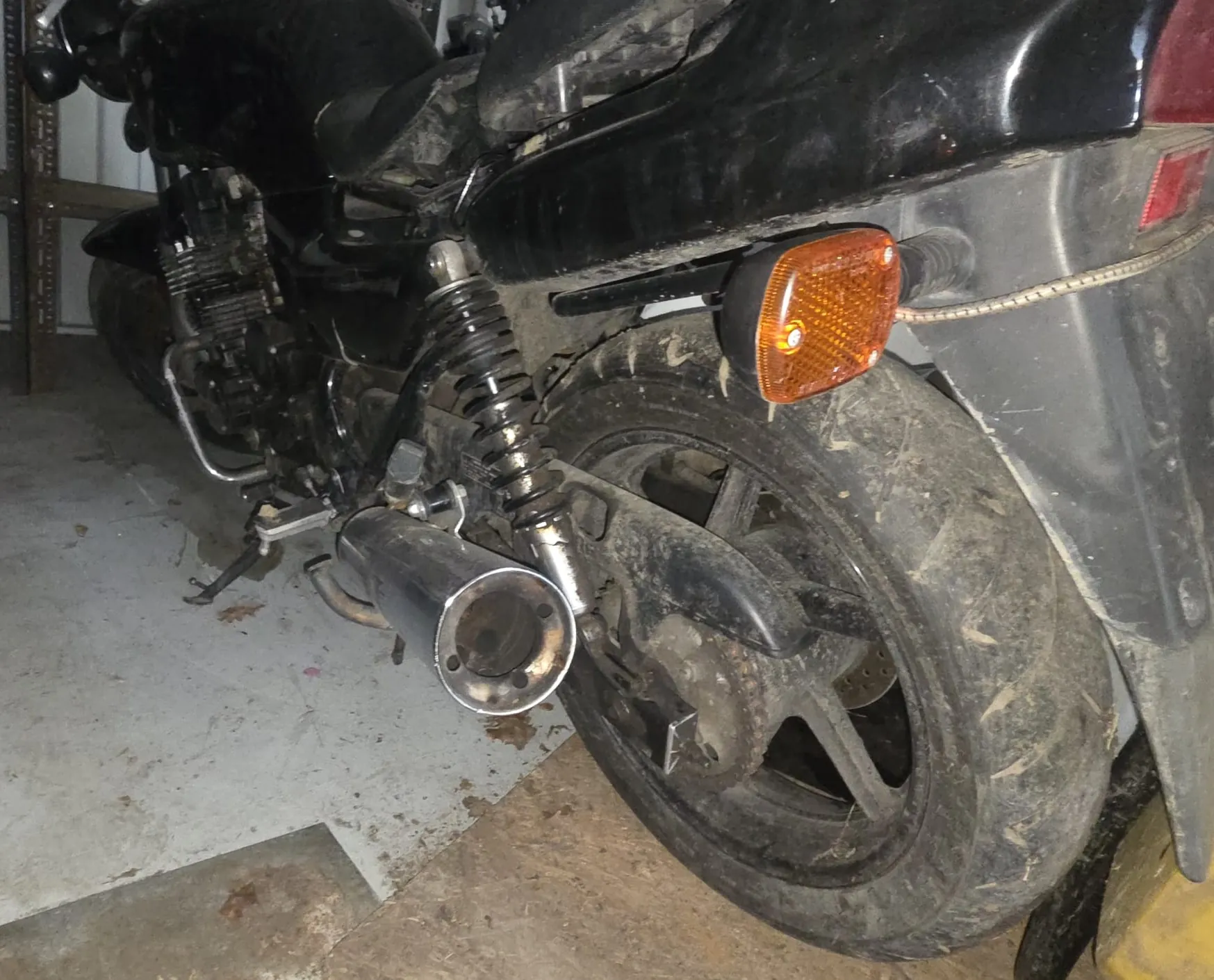 Zbliżenie na tylną część czarnego motocykla. Widoczne są brudne koło tylne z oponą, pomarańczowy kierunkowskaz, chromowany wydech oraz fragmenty mechanizmu zawieszenia. Motocykl stoi na betonowej podłodze z plamami, w tle metalowe konstrukcje. Brak czytelnego tekstu.