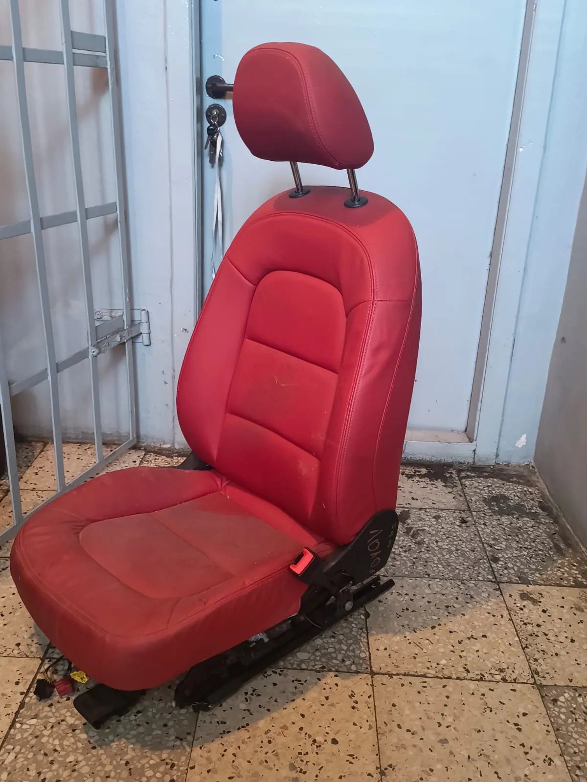 Czerwony, skórzany fotel samochodowy z zagłówkiem, ustawiony pionowo na jasnej, płytkowej podłodze przed jasnoniebieskimi drzwiami.