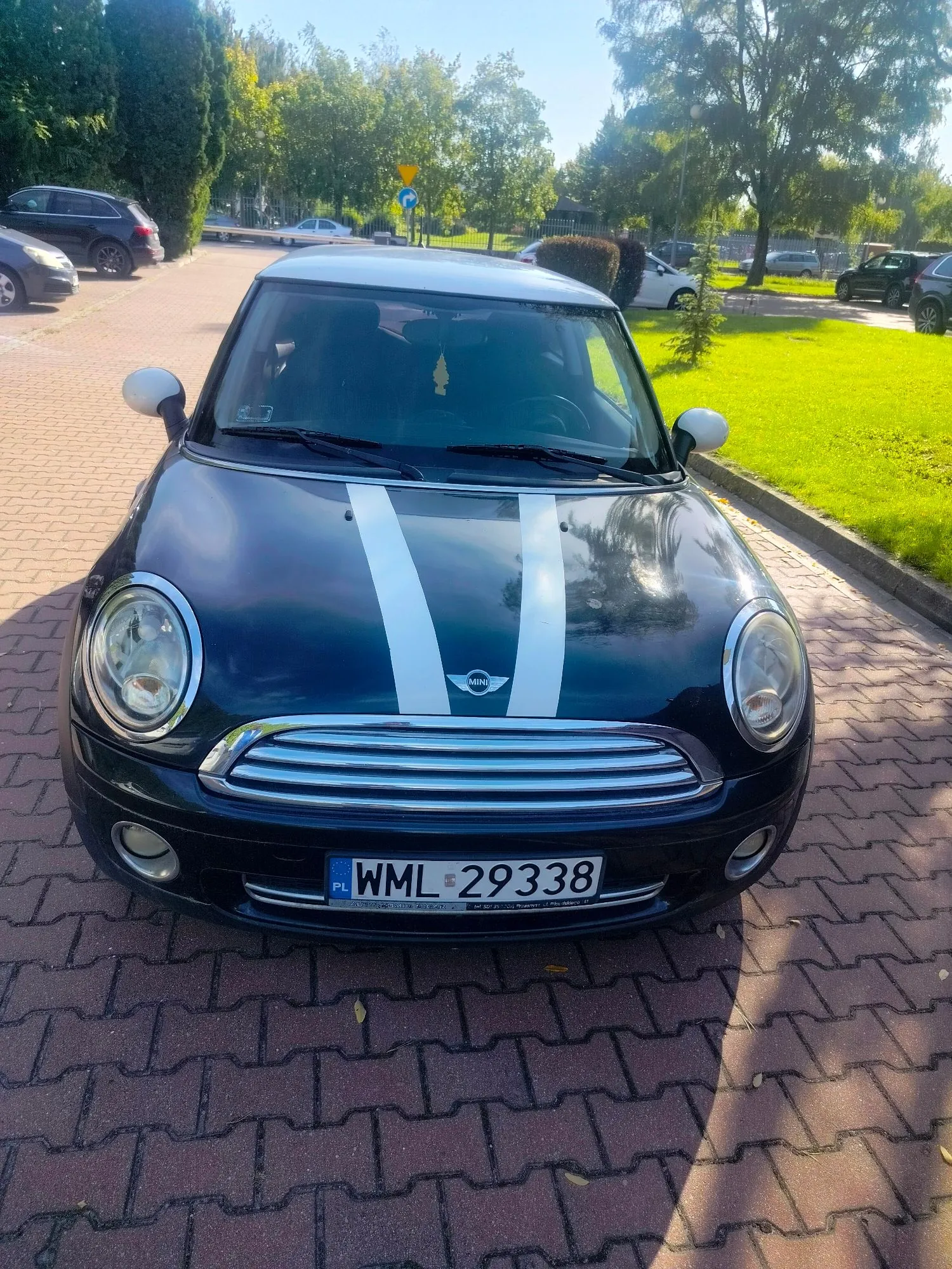 Czarny samochód marki Mini Cooper z białym dachem i dwoma białymi pasami na masce. Auto posiada polską tablicę rejestracyjną o numerze WML 29338. Samochód zaparkowany jest na kostce brukowej w słoneczny dzień, w tle widoczna jest trawa, drzewa oraz inne zaparkowane pojazdy.