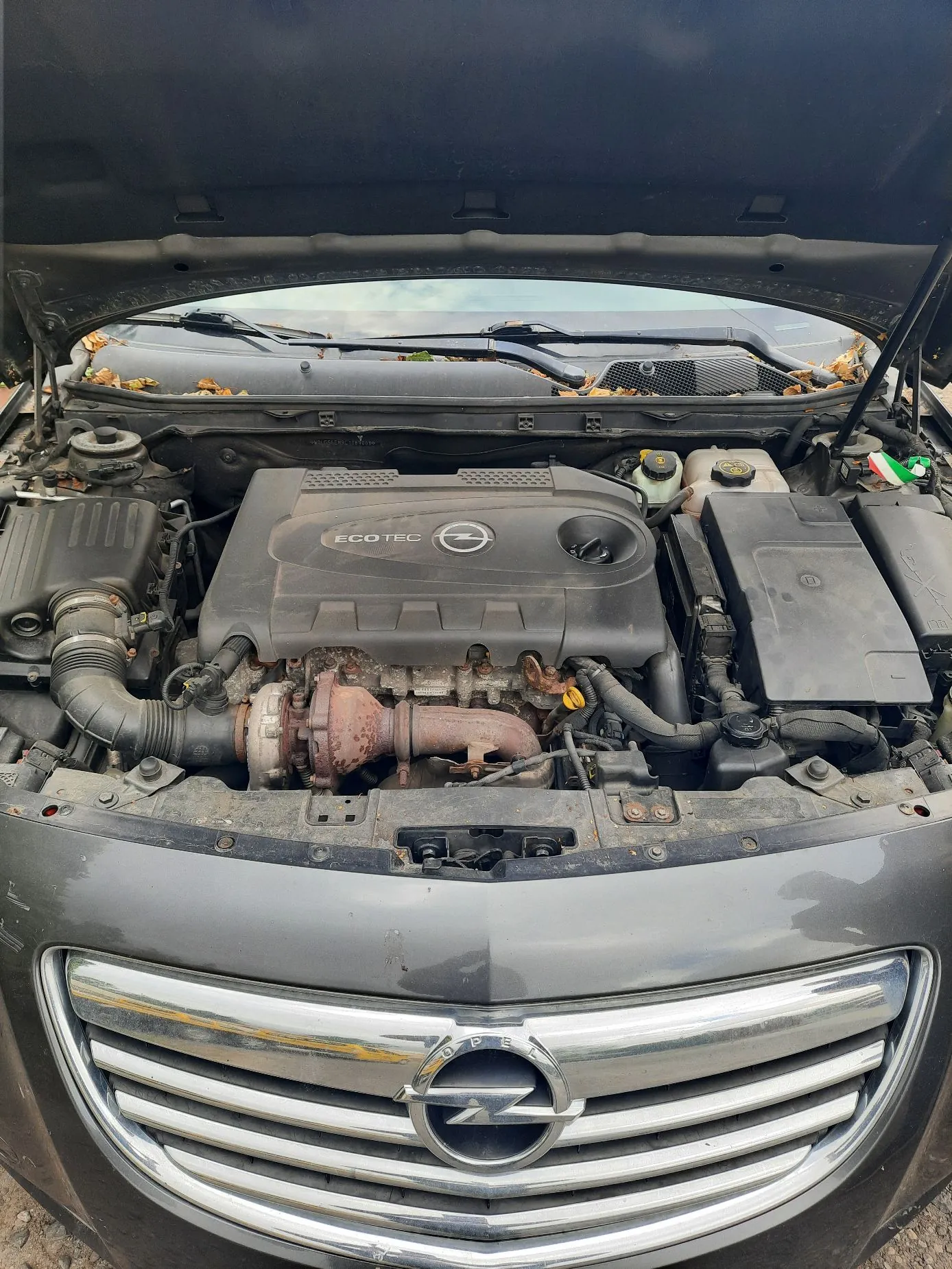 Widok z przodu na otwartą maskę ciemnoszarego samochodu marki Opel. Wewnątrz komory silnika widoczny jest silnik z plastikową osłoną z napisem 'ECOTEC' oraz logo Opela. Poniżej osłony znajduje się rdzawa turbosprężarka i układ wydechowy. Po prawej stronie widoczny jest czarny skrzynka bezpieczników i zbiorniki z płynami eksploatacyjnymi. Na dole zdjęcia znajduje się grill samochodu z chromowanymi listwami i dużym logo Opela w centrum.