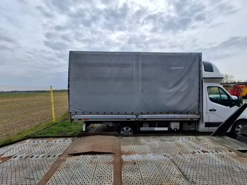 Biały samochód ciężarowy z przyczepą o szarej tkaninowej osłonie, zaparkowany na metalowej platformie z perforacjami. W tle żółte ogrodzenie, zielone pole, chmurne niebo oraz fragment czerwonego pojazdu po prawej stronie. Platforma posiada widoczne ślady korozji i uszkodzenia.