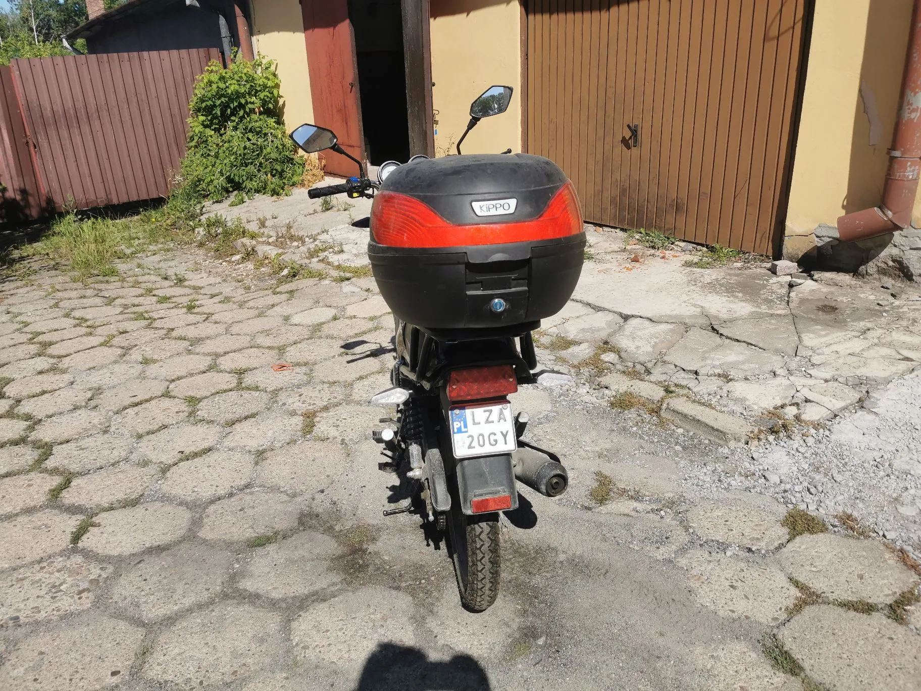 Widok z tyłu na czarny skuter zaparkowany na betonowym podjeździe z kostki brukowej. Skuter posiada czarny kufer z napisem 'KIPPO' i czerwonym odblaskiem. Widoczna jest polska tablica rejestracyjna o numerze 'LZA 20GY'. W tle znajduje się brązowy płot, zielony krzew oraz budynek z brązowymi drzwiami garażowymi.