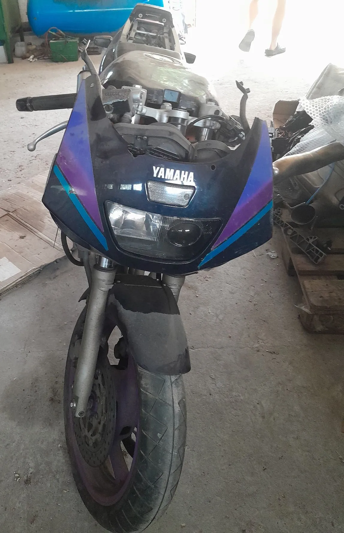 Widok z przodu na motocykl marki Yamaha w kolorze czarnym z fioletowymi i niebieskimi akcentami. Motocykl jest częściowo rozebrany, brakuje przedniej szyby. Na przedniej owiewce widnieje biały napis YAMAHA. Widoczny jest reflektor, przednie koło z oponą oraz widelec. Motocykl stoi w garażu lub warsztacie na betonowej podłodze.