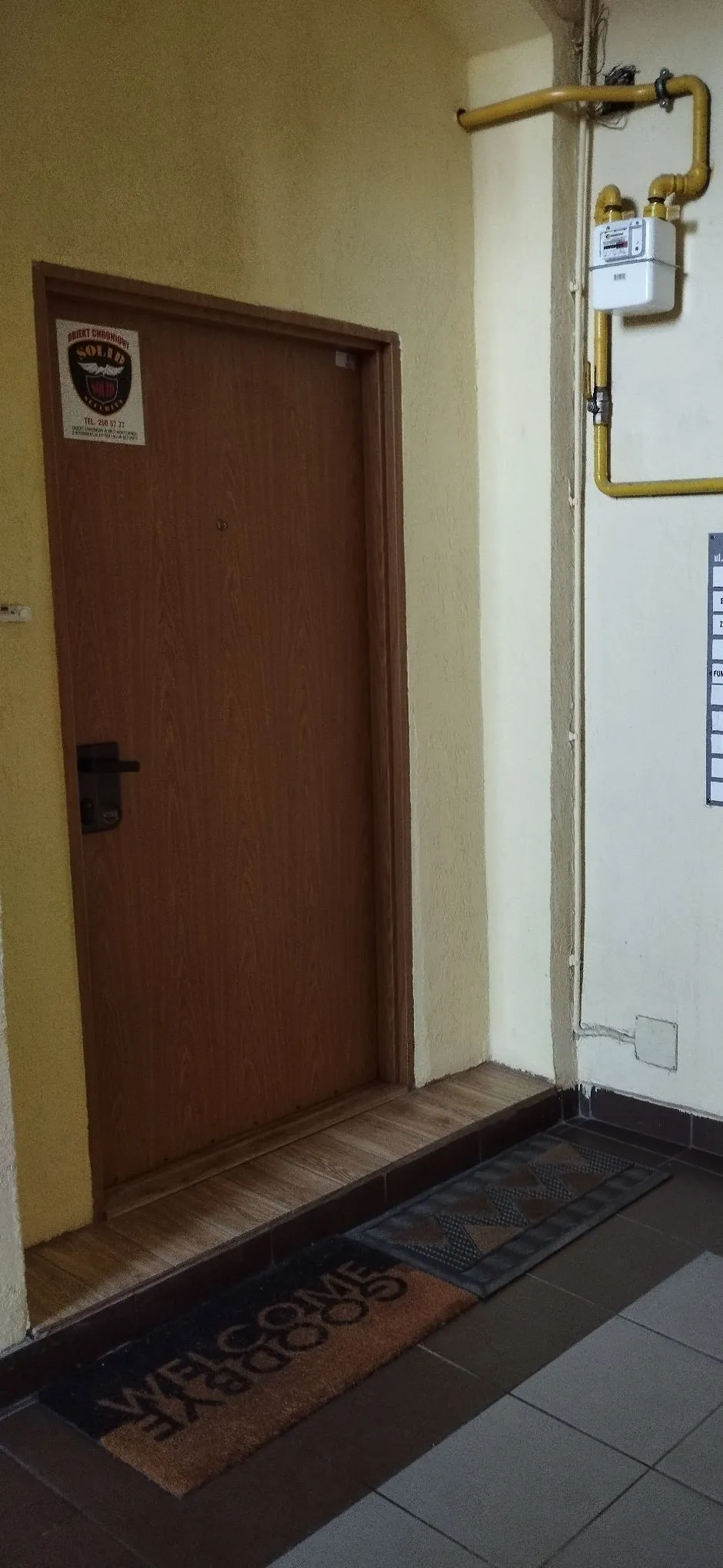 Zdjęcie korytarza z brązowymi drzwiami z drewna. Na drzwiach znajduje się naklejka z napisem 'BUDYNKI SAMODZIELNE SOLID' oraz numerem telefonu 'Tel. 226 52 27'. Obok drzwi, na białej ścianie, biegnie żółta rura gazowa z zamontowanym białym licznikiem. Przed drzwiami na podłodze leżą dwie wycieraczki: jedna ciemna z wzorem geometrycznym, a druga brązowa z napisem 'WELCOME' u góry i 'GOODBYE' u dołu.