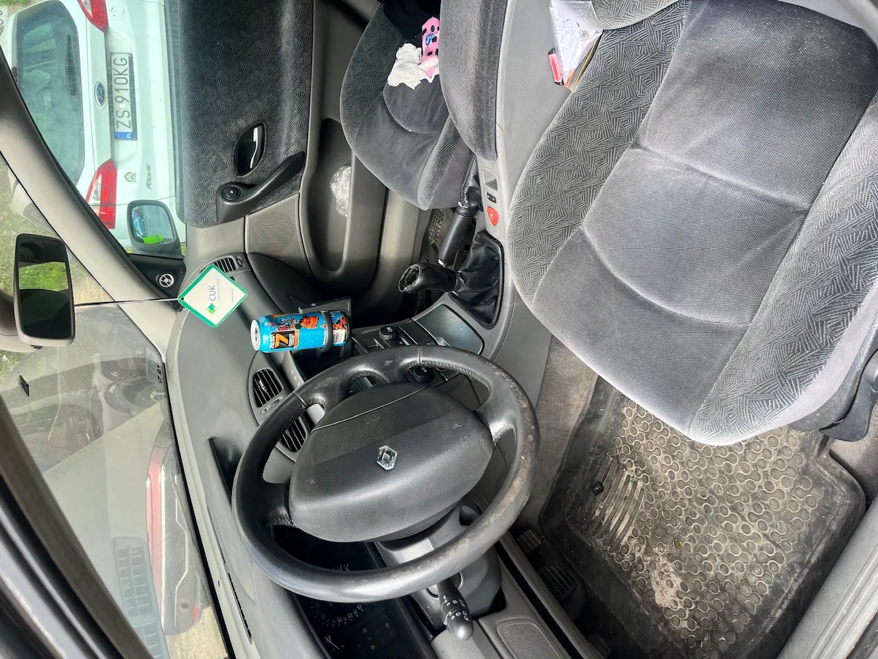 Wnętrze samochodu widziane z perspektywy kierowcy. Widoczna jest czarna kierownica z logo Renault, szary fotel pasażera oraz deska rozdzielcza. Na desce rozdzielczej leży niebieska puszka napoju energetycznego oraz mała, zielono-biała karteczka. Przez przednią szybę i okno widać fragment białego samochodu z tablicą rejestracyjną ZS 910KG.
