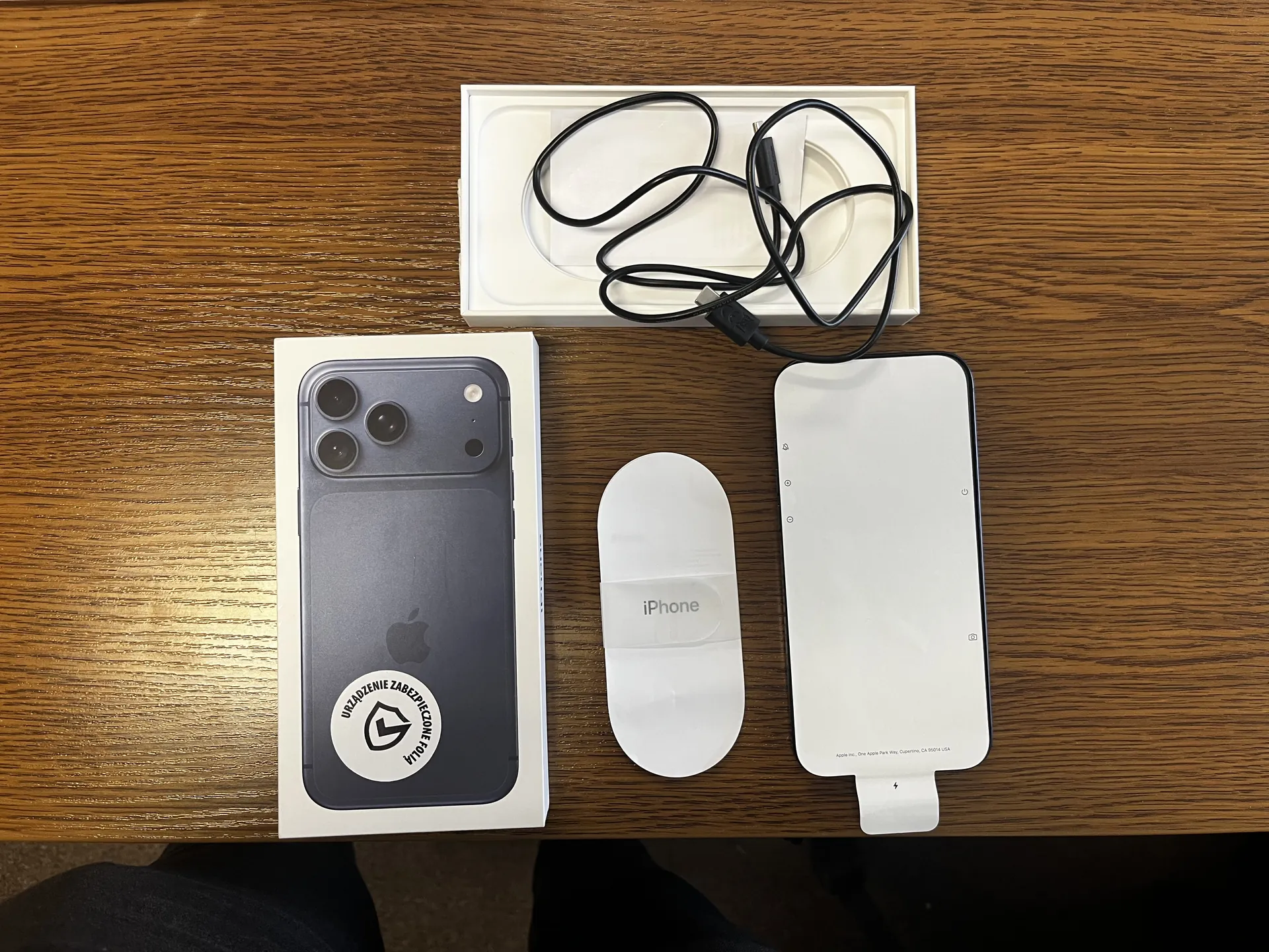 Zdjęcie przedstawia drewniany stół z ułożonymi na nim przedmiotami: pudełko z czarnym iPhone'em (widoczny logo Apple i naklejka z napisem 'URZĄDZENIE ZABEZPIECZONE POLA' oraz symbolem tarczy), białą owalną podkładkę z napisem 'iPhone', białą prostokątną folię ochronną z małym tekstem na dole oraz otwarte białe pudełko z czarnym przewodem USB. Wszystkie elementy są ułożone równolegle do krawędzi zdjęcia.