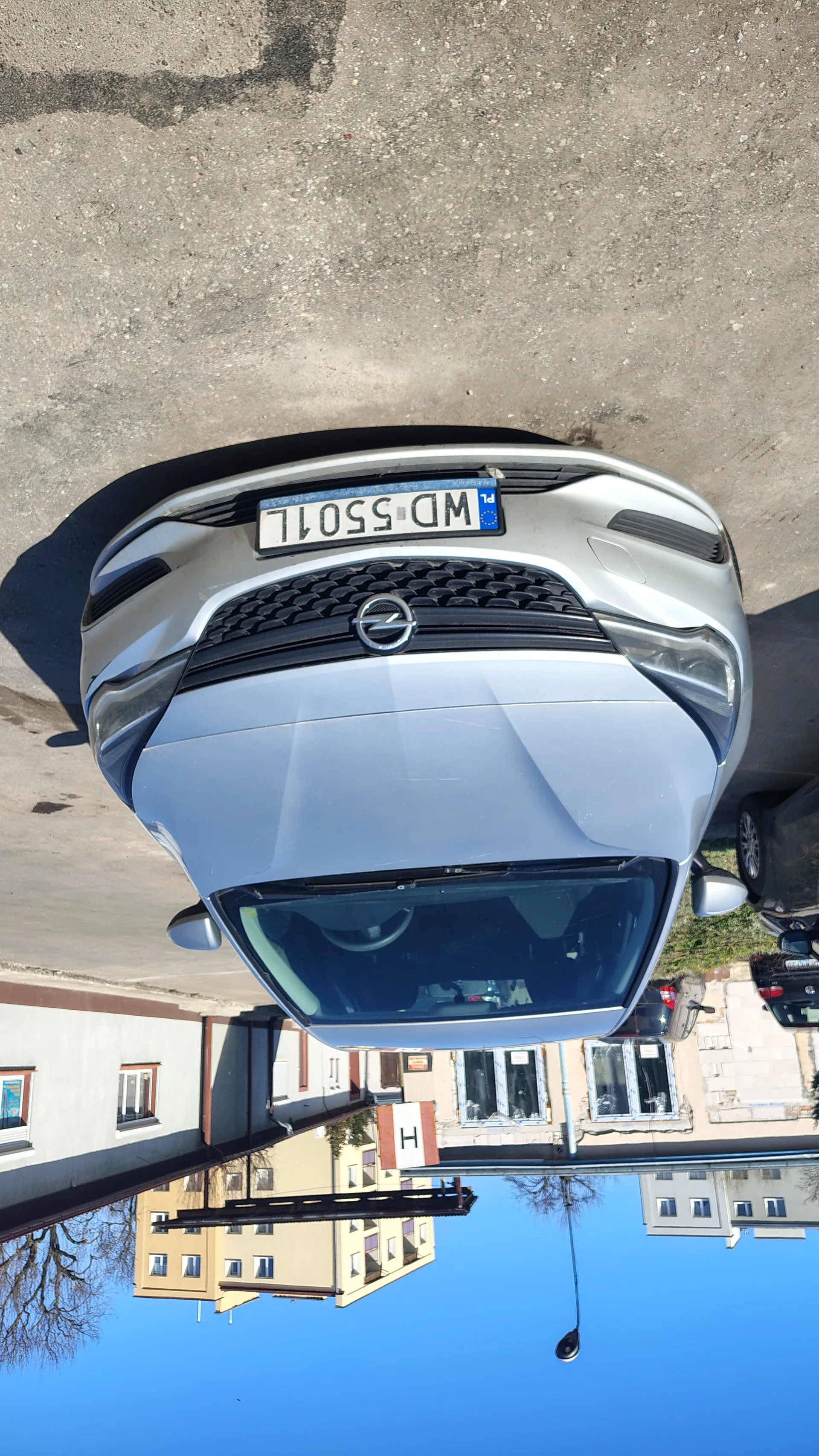 Srebrny samochód marki Opel zaparkowany na asfaltowej powierzchni. Widoczna tablica rejestracyjna z napisem "WD 5501L" oraz oznaczeniem "PL". W tle budynki mieszkalne, inne zaparkowane samochody, latarnia uliczna i bezchmurne niebo. Na jednym z budynków widoczny znak z literą "I".