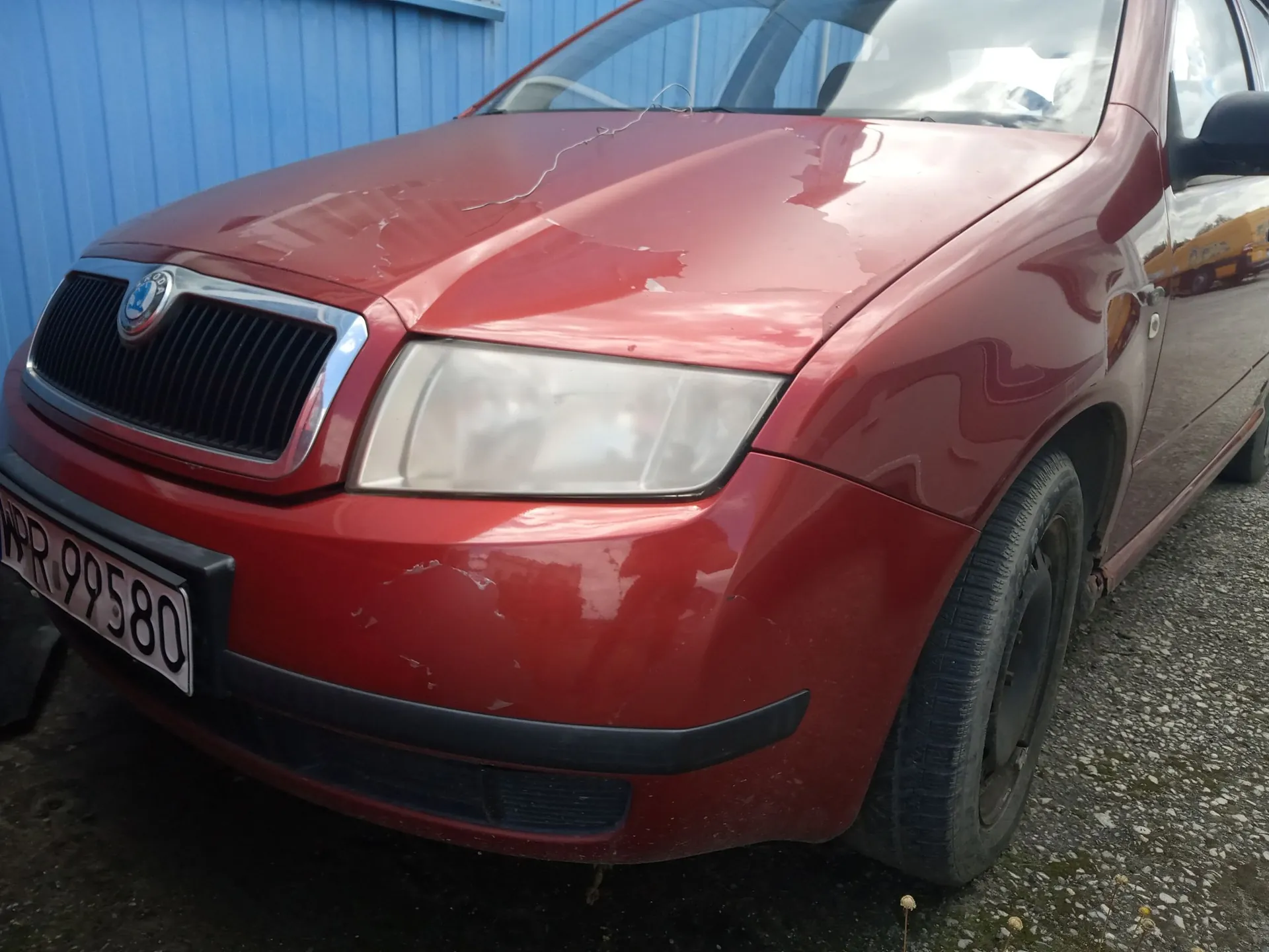 Zbliżenie na przód czerwonego samochodu marki Skoda. Widoczna jest maska z licznymi wgnieceniami i zarysowaniami, zmatowiony reflektor prawy oraz czarny grill z logo marki. Na zderzaku znajduje się tablica rejestracyjna z widocznym fragmentem tekstu 'PR 99580'. Samochód stoi na szutrowym podłożu na tle niebieskiego ogrodzenia.