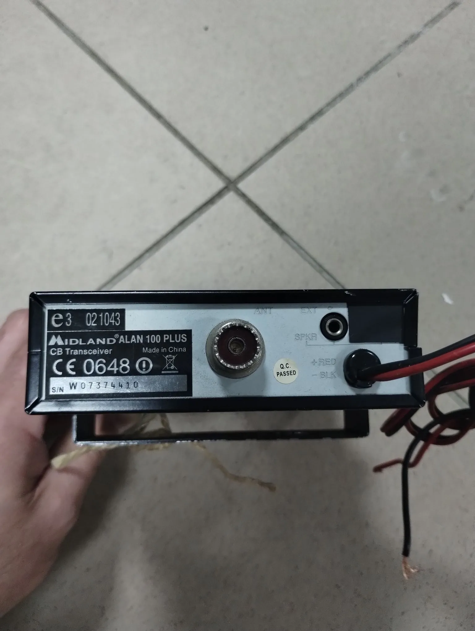 Zdjęcie tylnego panelu radiostacji CB Midland Alan 100 Plus. Na srebrnej naklejce widnieje napis 'MIDLAND ALAN 100 PLUS CB Transceiver', 'Made in China', oznaczenie 'CE 0648' oraz numer seryjny 'S/N W07374410'. Na panelu znajduje się gniazdo antenowe (ANT), gniazdo głośnika (SPKR) oraz dwa przewody zasilające w kolorach czerwonym i czarnym z opisami '+ RED' i '- BLK'.
