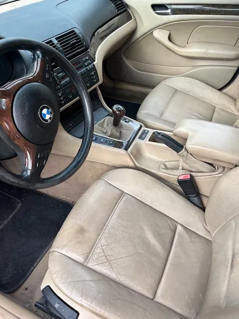 Wnętrze samochodu marki BMW z beżową skórzaną tapicerką. Widoczna jest kierownica z logo BMW, konsola środkowa z lewarkiem zmiany biegów, hamulec ręczny oraz dwa przednie fotele. Deska rozdzielcza jest w kolorze czarnym z drewnianymi elementami wykończenia.