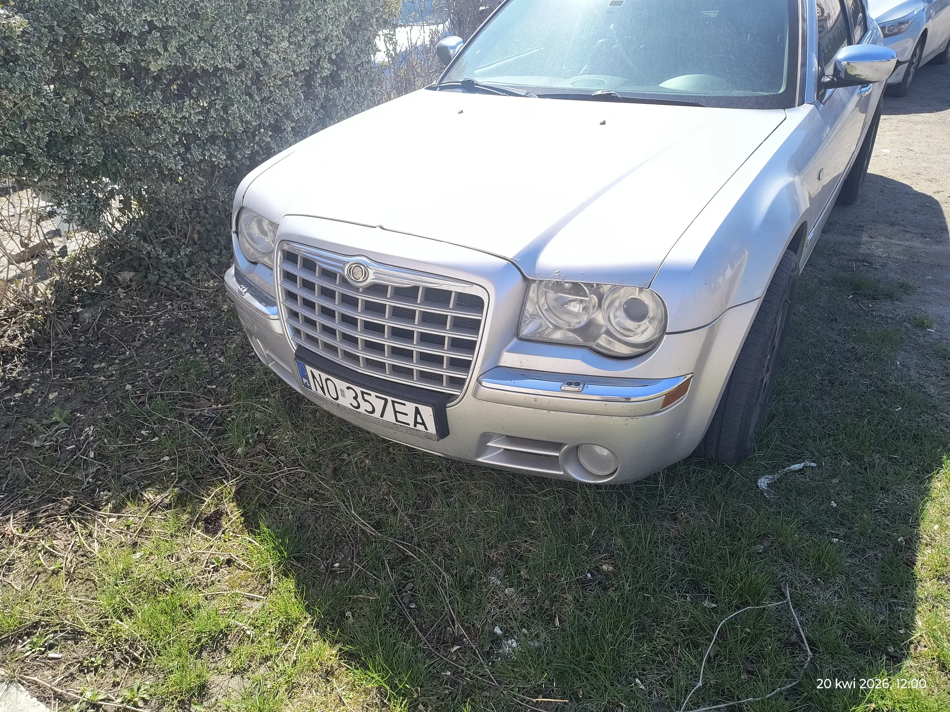Przód srebrnego samochodu marki Chrysler z tablicą rejestracyjną NO 357EA, zaparkowanego na trawie. W tle krzew, fragment drogi i inny samochód. W prawym dolnym rogu widoczny znaczek czasu: 20 kwi 2026, 12:00