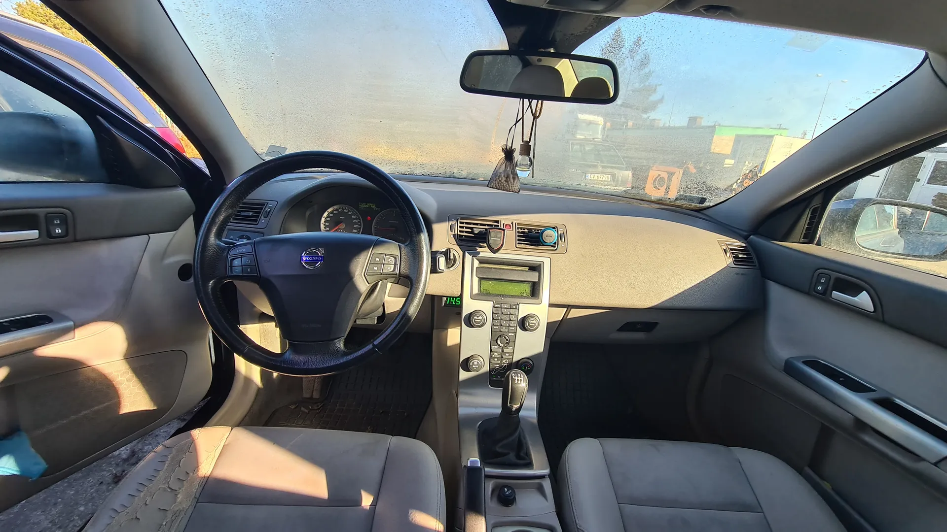 Wnętrze samochodu Volvo z widokiem na kierownicę z logo marki, deski rozdzielczej z licznikami, centralną konsolą z ekranem i przyciskami oraz dźwignią zmiany biegów. Widoczne są szare fotele, lusterko wsteczne z wiszącym elementem oraz zewnętrzny widok przez przednią szybę na budynki i inne pojazdy. Kierownica znajduje się po lewej stronie, a tekst na licznikach jest czytelny i ustawiony pionowo.