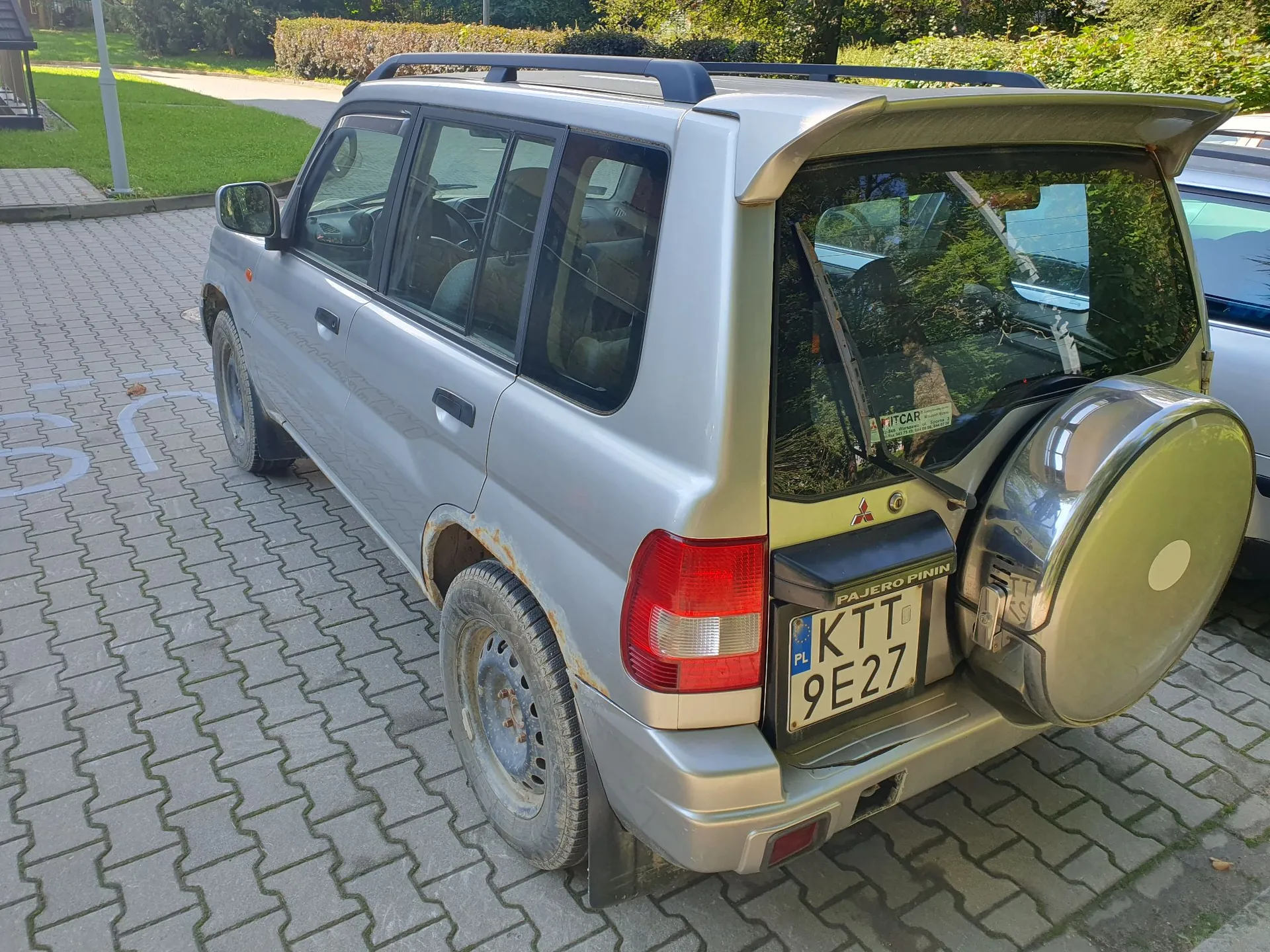 Srebrny samochód terenowy Mitsubishi Pajero Pinin zaparkowany na kostce brukowej. Pojazd jest widoczny od tyłu i z boku, posiada koło zapasowe zamontowane na tylnych drzwiach oraz tablicę rejestracyjną o numerze KTT 9E27. Na nadkolu widoczne są ślady rdzy.