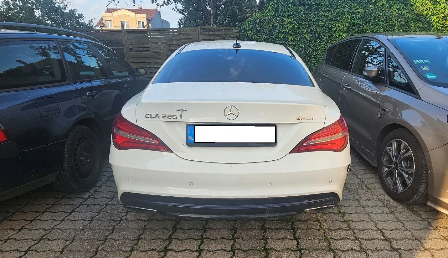 Widok z tyłu białego samochodu marki Mercedes-Benz model CLA 220 T 4Matic, zaparkowanego między ciemnoniebieskim kombi po lewej a brązowym samochodem po prawej. Pojazd posiada polską tablicę rejestracyjną, która jest zakryta białym prostokątem. Samochody stoją na kostce brukowej przed drewnianym płotem i zieloną ścianą z roślinności.