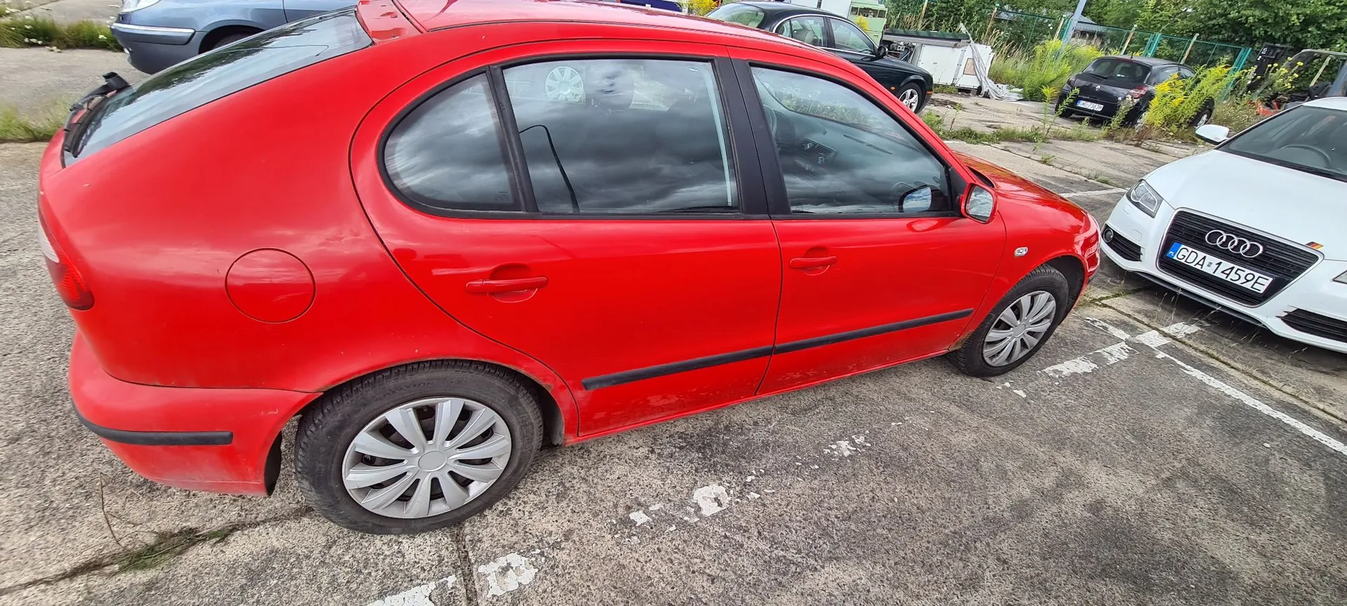 Czerwony samochód osobowy typu hatchback zaparkowany na betonowym placu. Widok z boku, od strony pasażera. W tle widoczne są inne zaparkowane samochody, w tym biały Audi z tablicą rejestracyjną GDA 1459E oraz ciemne auto. W oddali widać zielone krzewy i drzewa.
